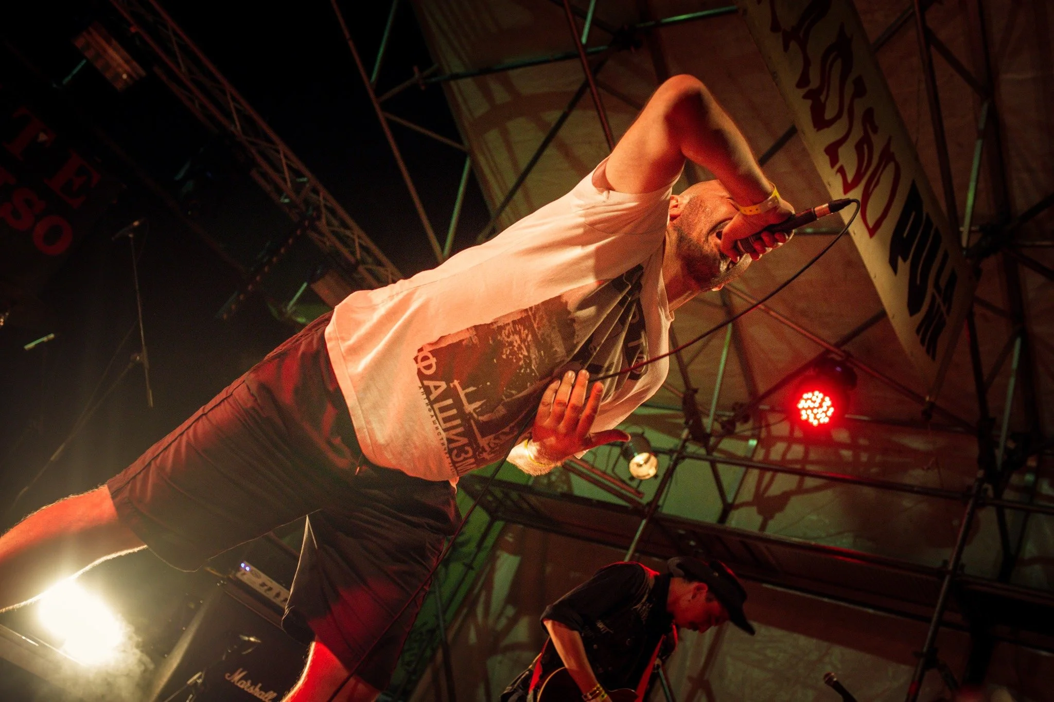 A male singer performing on stage with a microphone, wearing a white T-shirt and dark shorts, in a concert setting with stage lights and equipment.