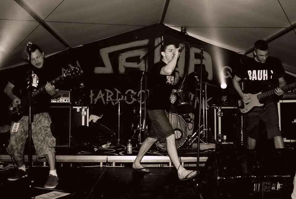 Band performing live on stage with three guitarists and a vocalist, in a tented venue with musical equipment and lighting.