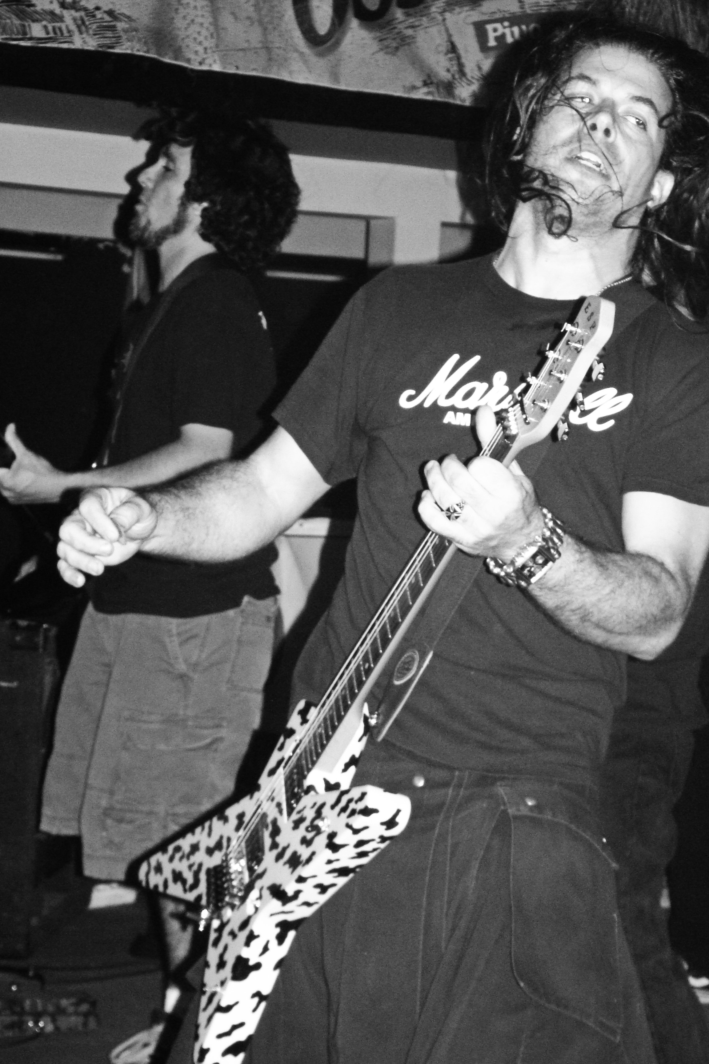 Two men playing electric guitars, one in focus with long wavy hair and a Mohawk t-shirt, the other in the background with curly hair, in a room with posters on the wall.