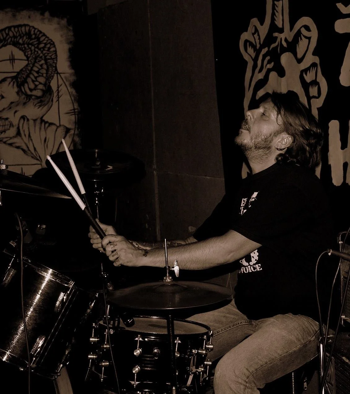 A man with long hair and tattoos playing drums in a dimly lit room with dark walls, featuring artwork and designs.