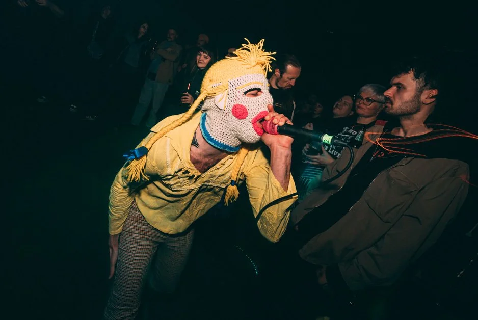 Person wearing a colorful, sock puppet-like mask with yellow yarn hair singing into a microphone at a concert, with an audience watching.