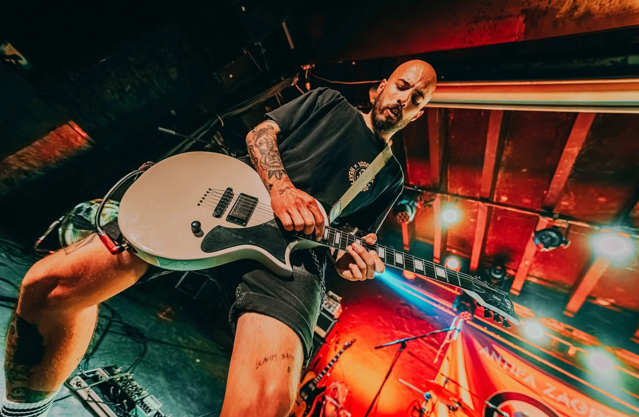 A man with tattoos playing a white electric guitar on stage in a dimly lit venue with red and orange lighting.