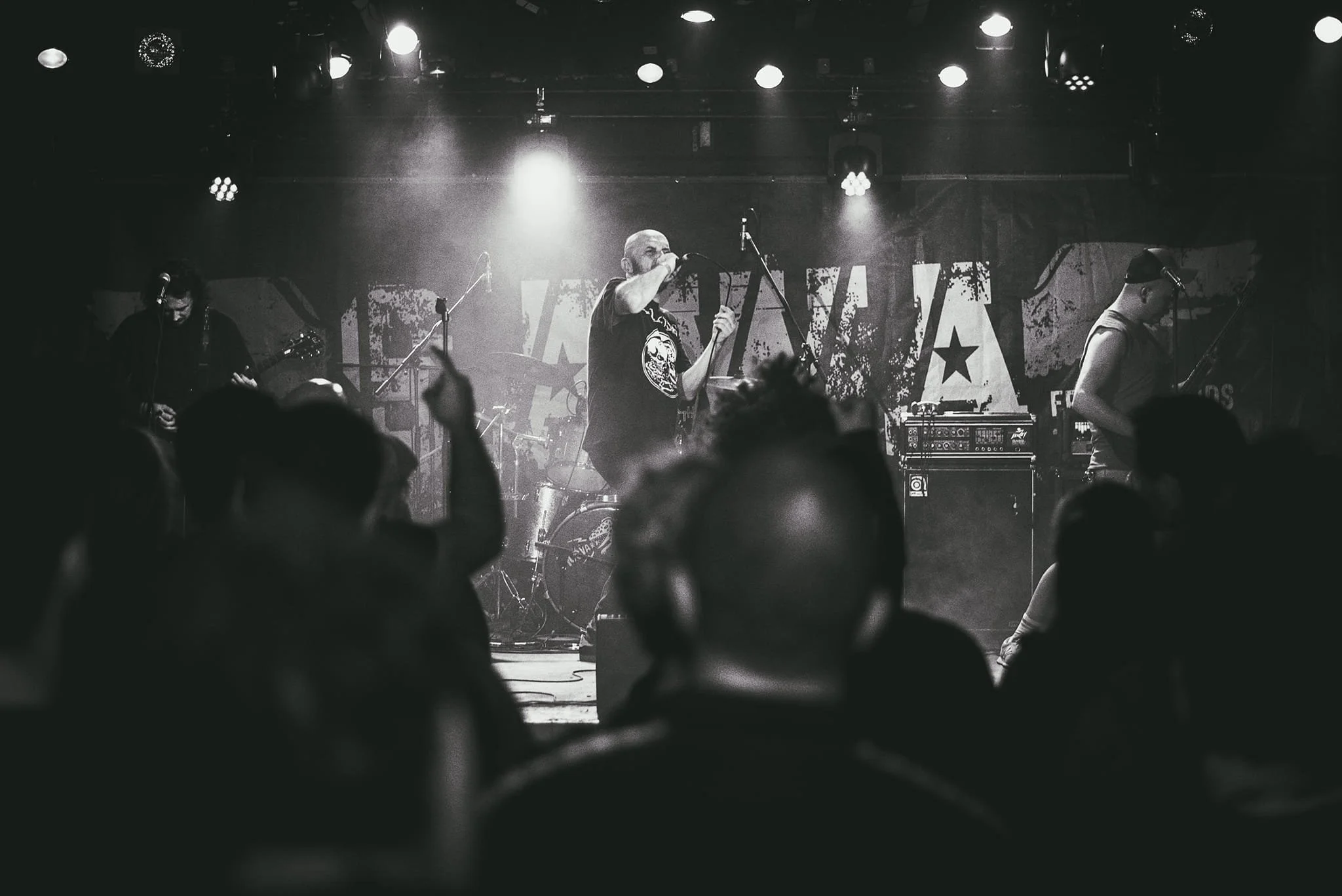 A live music concert with a band performing on stage. The lead singer is holding a microphone and appears to be singing. There are two other band members playing guitar, and various musical equipment on stage. Audience members are in the foreground w