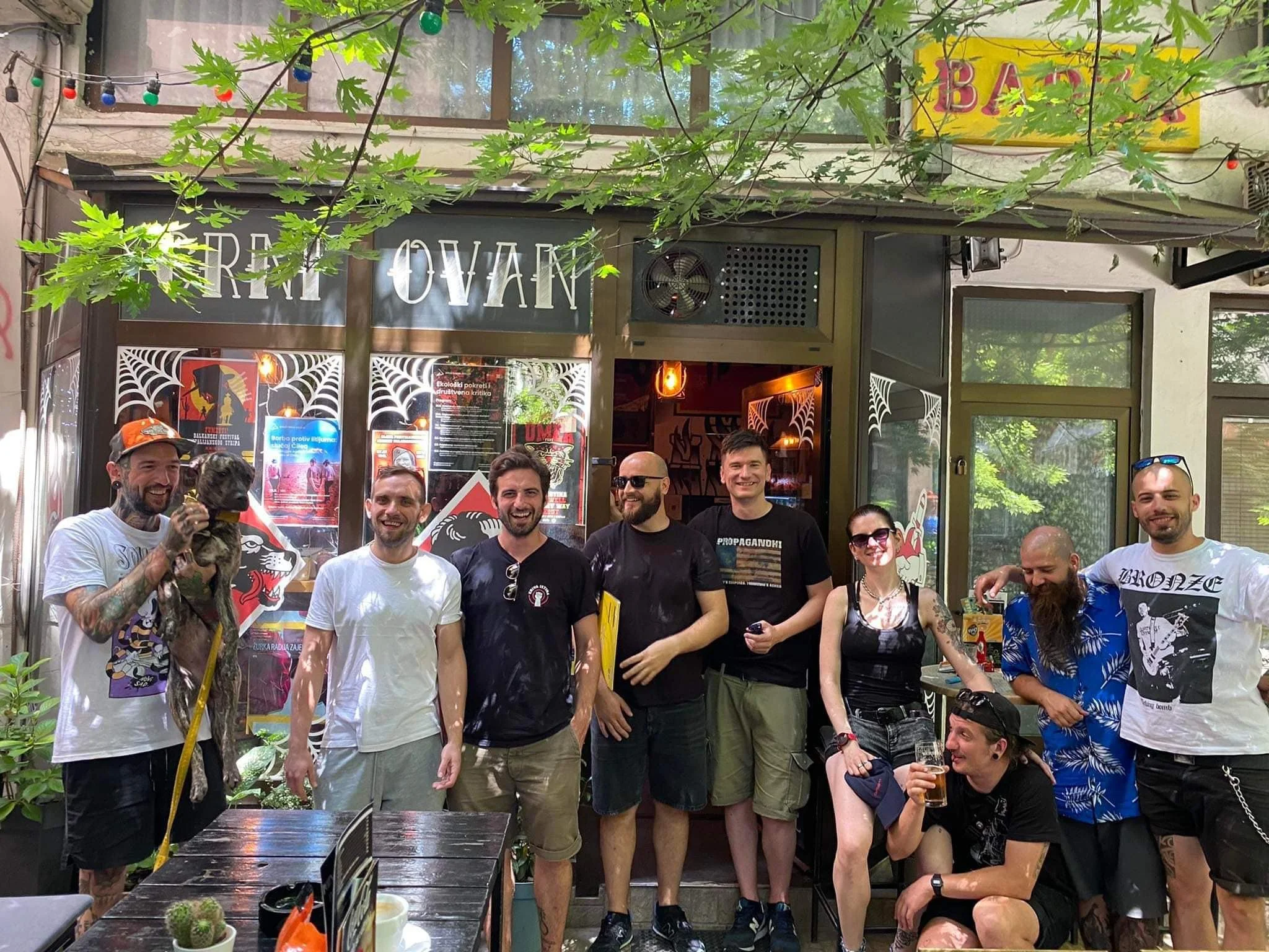 A group of people smiling and gathered outside a restaurant or bar, with some holding drinks. There are men and women of various ages, and the setting appears to be casual and lively. The background shows a sign, some posters, greenery, and string li
