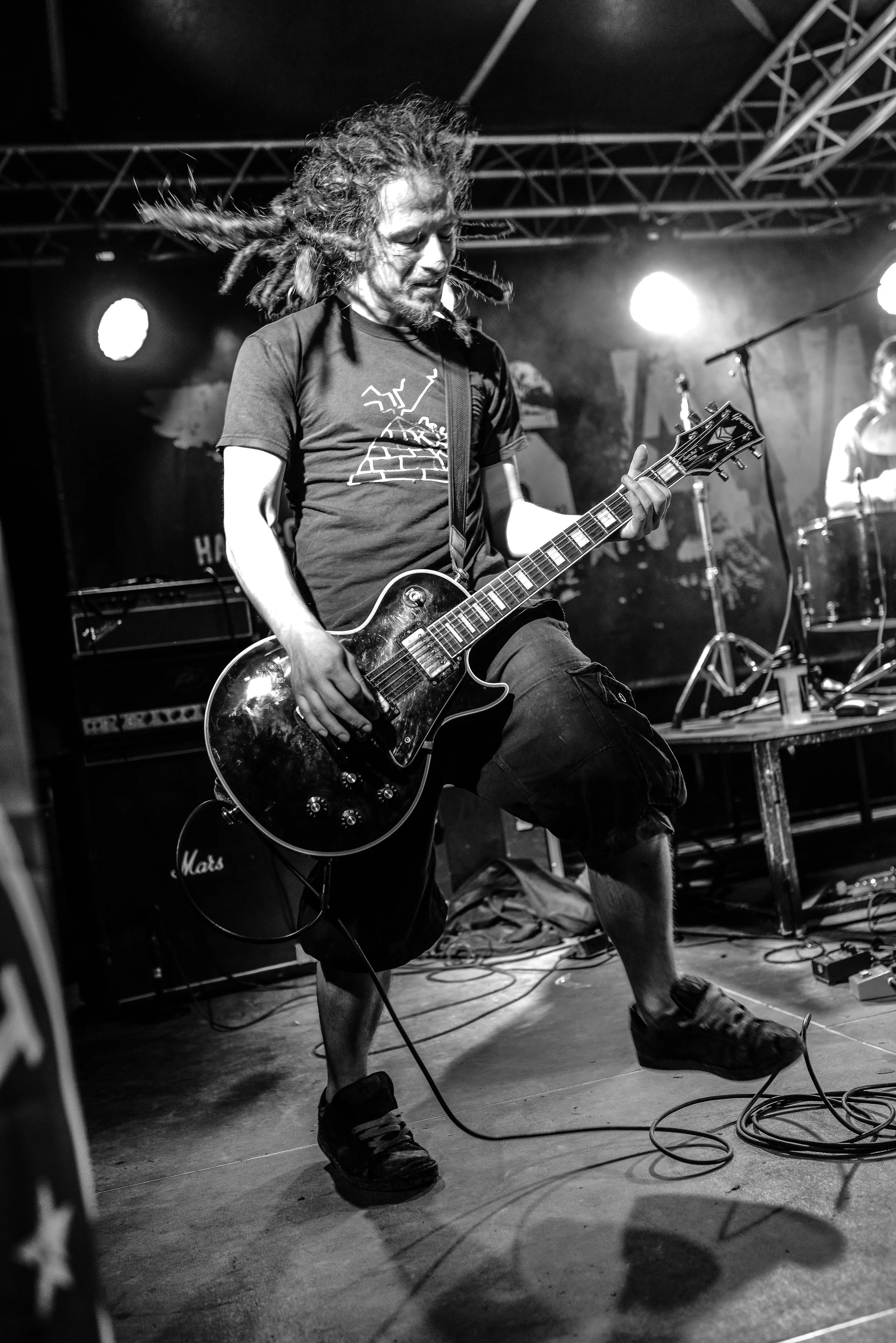 A musician with dreadlocks playing an electric guitar on stage during a live performance, with stage lighting and equipment visible in the background and shadows cast on the floor.