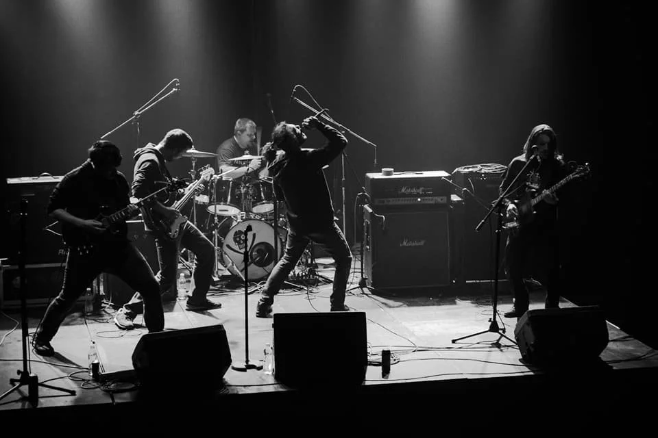 A band performing live on stage with five musicians playing guitars, drums, and singing, surrounded by microphones and amplifiers, in a dark setting.