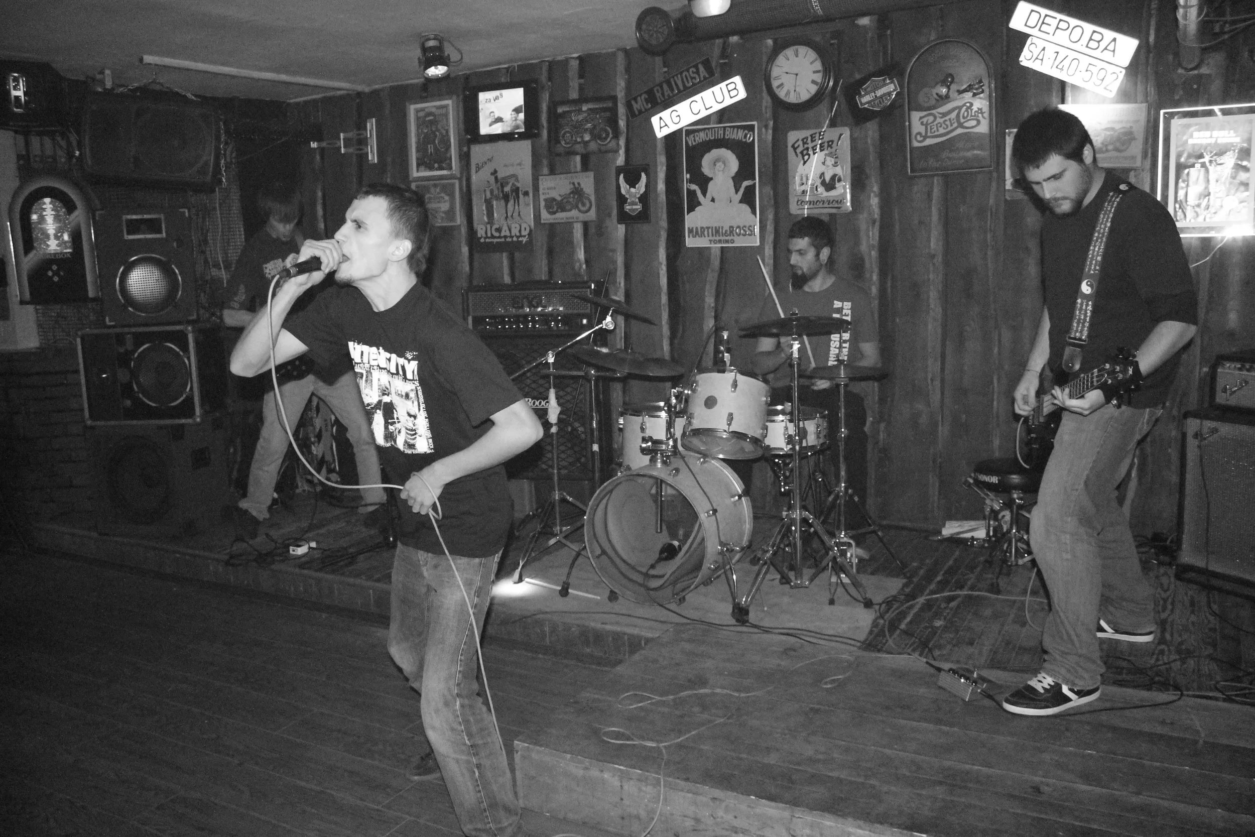 A band performing in a bar with three male musicians on stage. One is singing into a microphone, the second is playing drums, and the third is playing an electric guitar. The background has various posters and signs on the wooden wall, including a cl