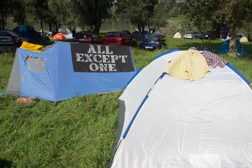 Tent with a sign that says 'ALL EXCEPT ONE' in a grassy outdoor camping area, with other tents and parked cars in the background.