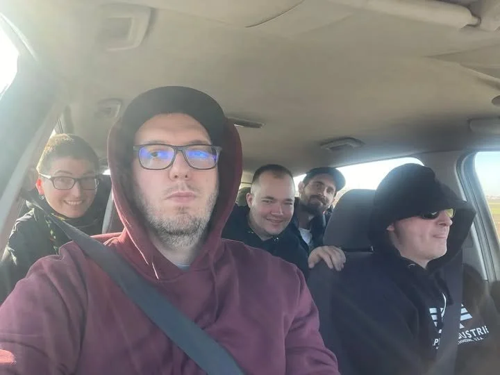 Five men sitting in a car, all wearing hoodies or jackets, with the person in the front wearing glasses and a maroon hoodie, taking a selfie.