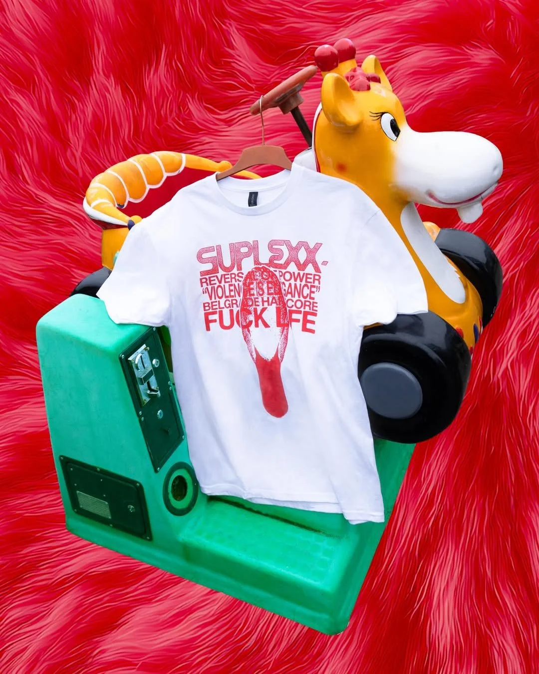 A colorful amusement park ride shaped like a giraffe with a white t-shirt hanging on a hanger in front of it. The background has red, fluffy textured material, and there is a teal speaker or boombox on the left side.