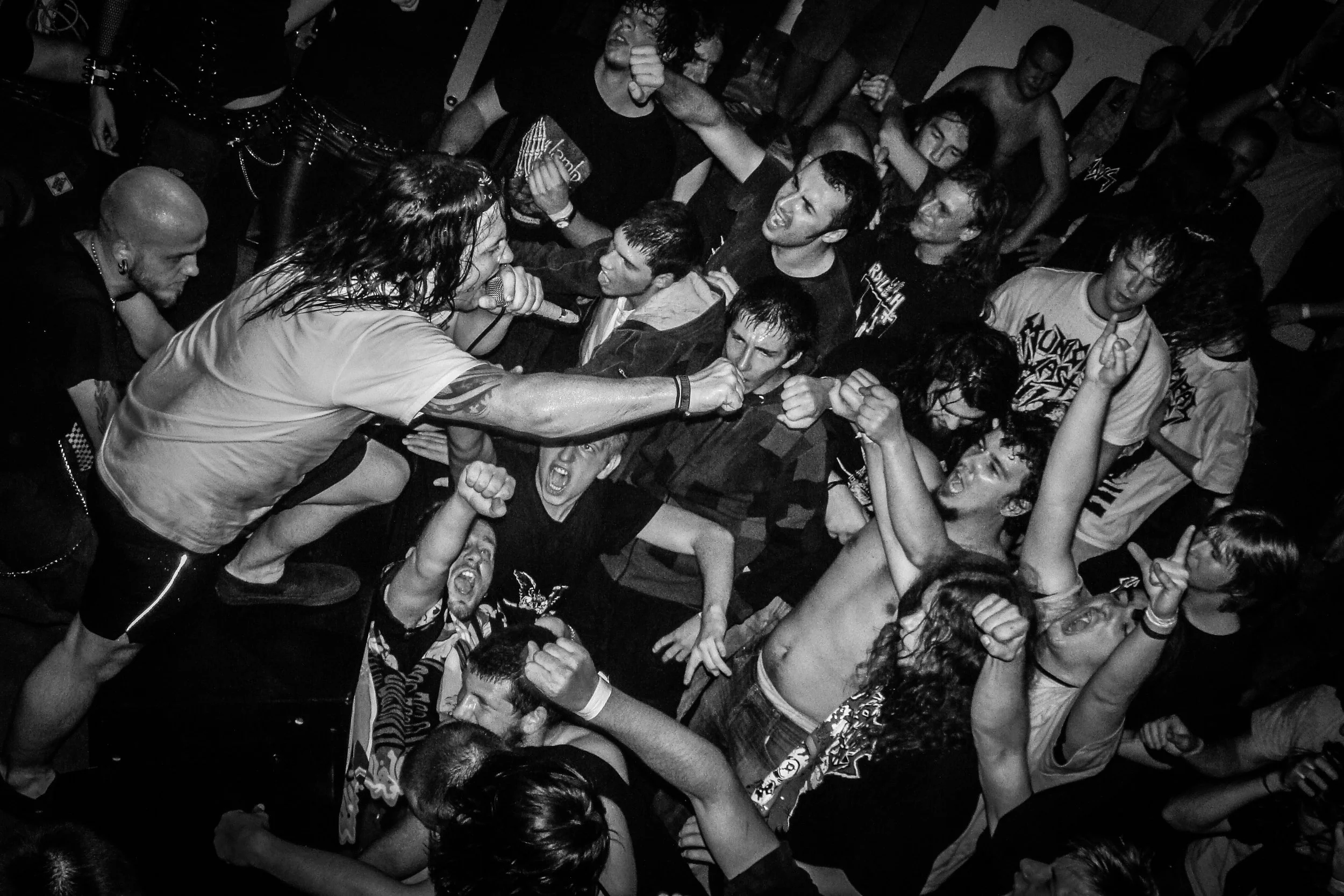 A black-and-white photo of a crowded concert or music event with people singing, shouting, and engaging intensely with the performer on stage. The scene displays energetic emotions and a sense of chaos and excitement. Band name: Municipal Waste