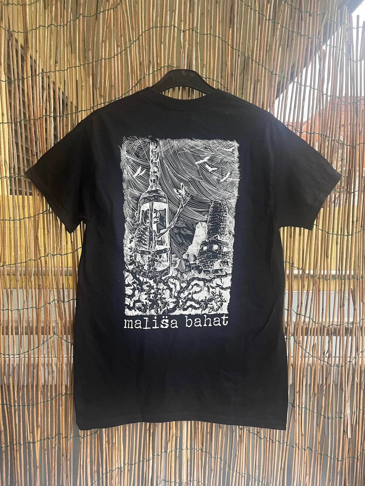 Black T-shirt with a white graphic design depicting a Native American figure, cacti, and a desert landscape, with the text "malisa bahat" underneath, hanging on a black hanger in front of a bamboo screen.