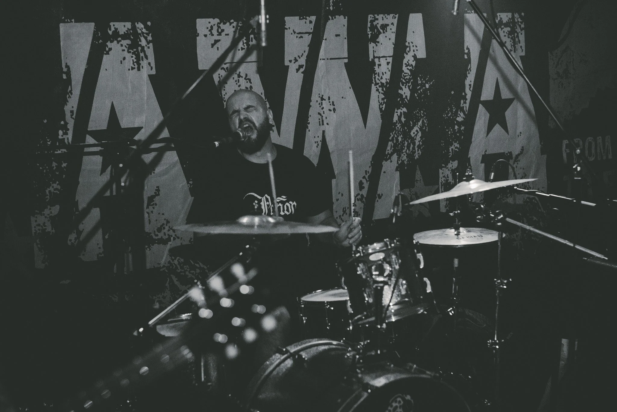 A musician passionately singing and playing drums during a live performance with a large banner behind him.
