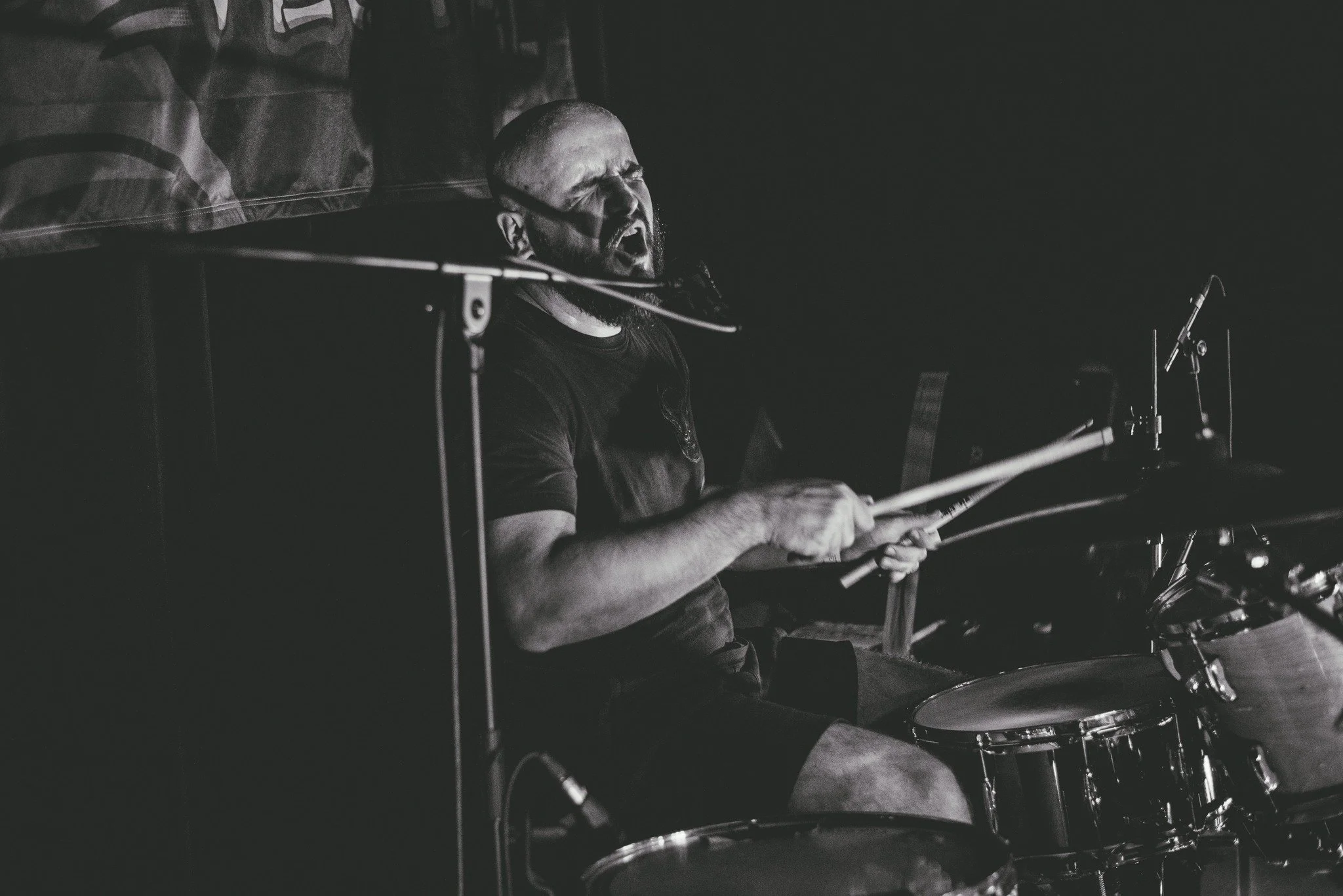 A man with a beard playing the drums passionately, with his eyes closed and mouth open, in a dark room.