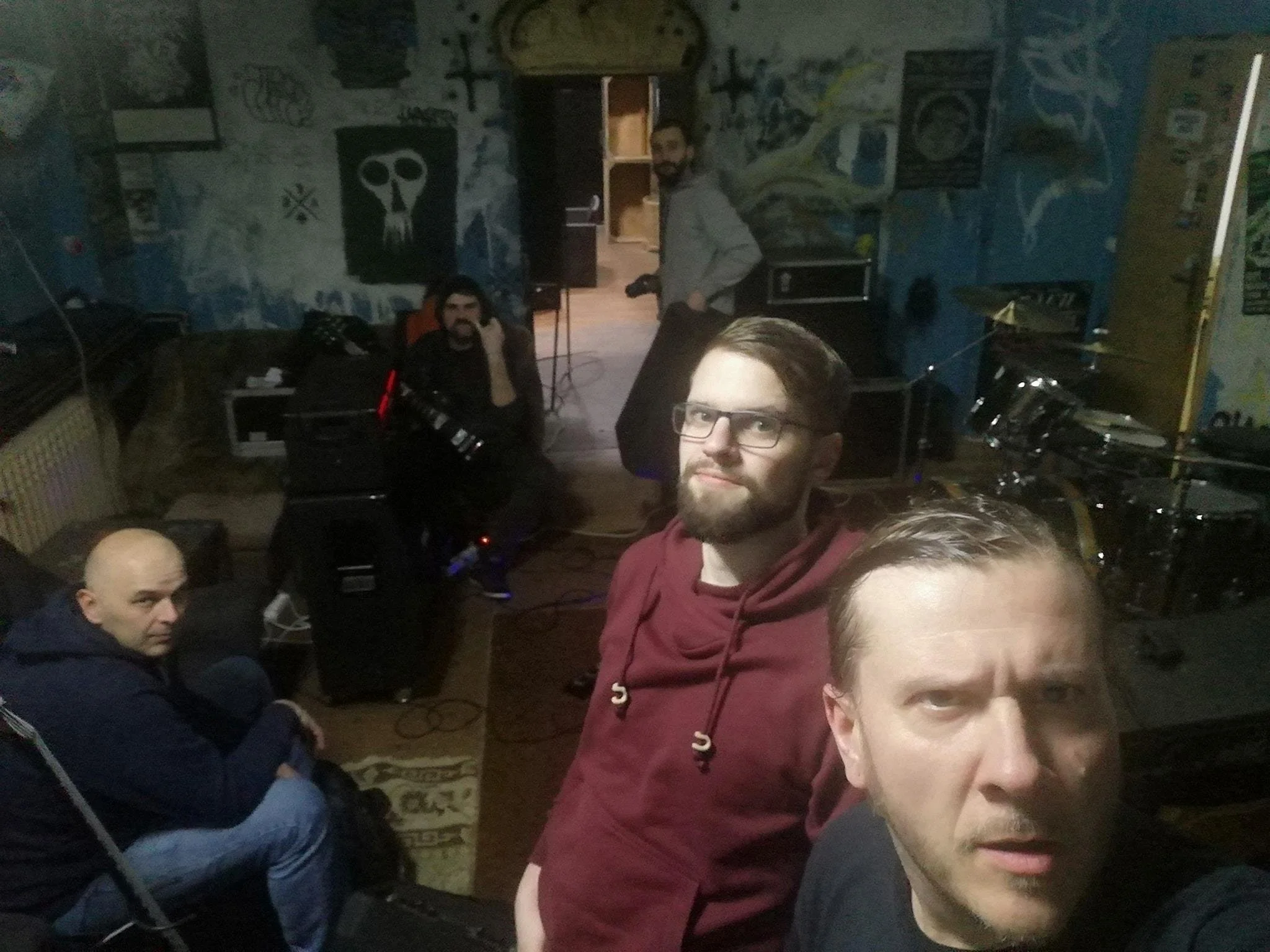 People at a dimly lit indoor gathering, some sitting and some standing, with graffiti-covered walls and music equipment in the background.