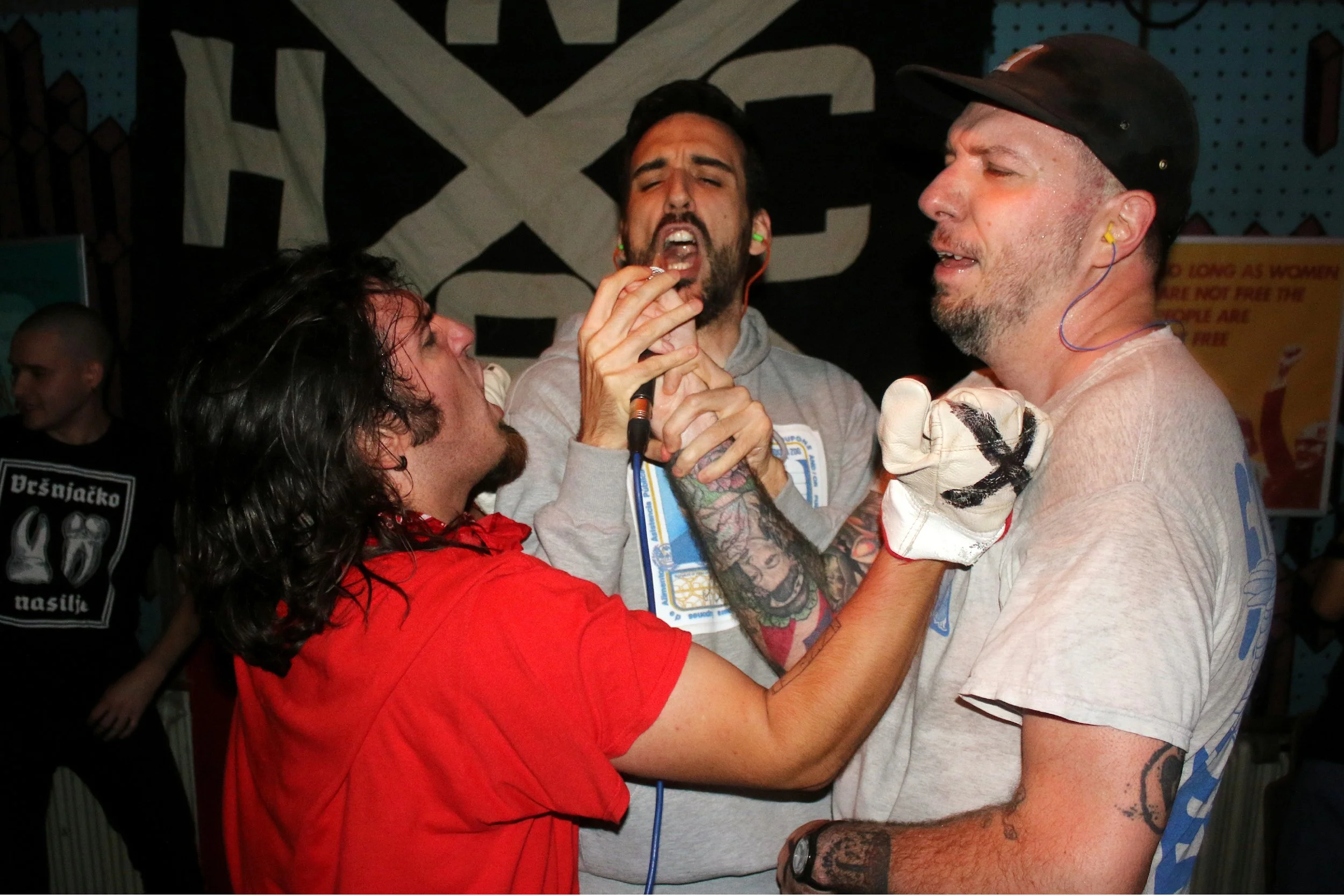 A woman in a red shirt is wrestling with a man with tattoos and a beard, who is wearing a gray hoodie and a black cap. They appear to be at a lively event with a large black and white banner in the background, and other people can be seen in the back