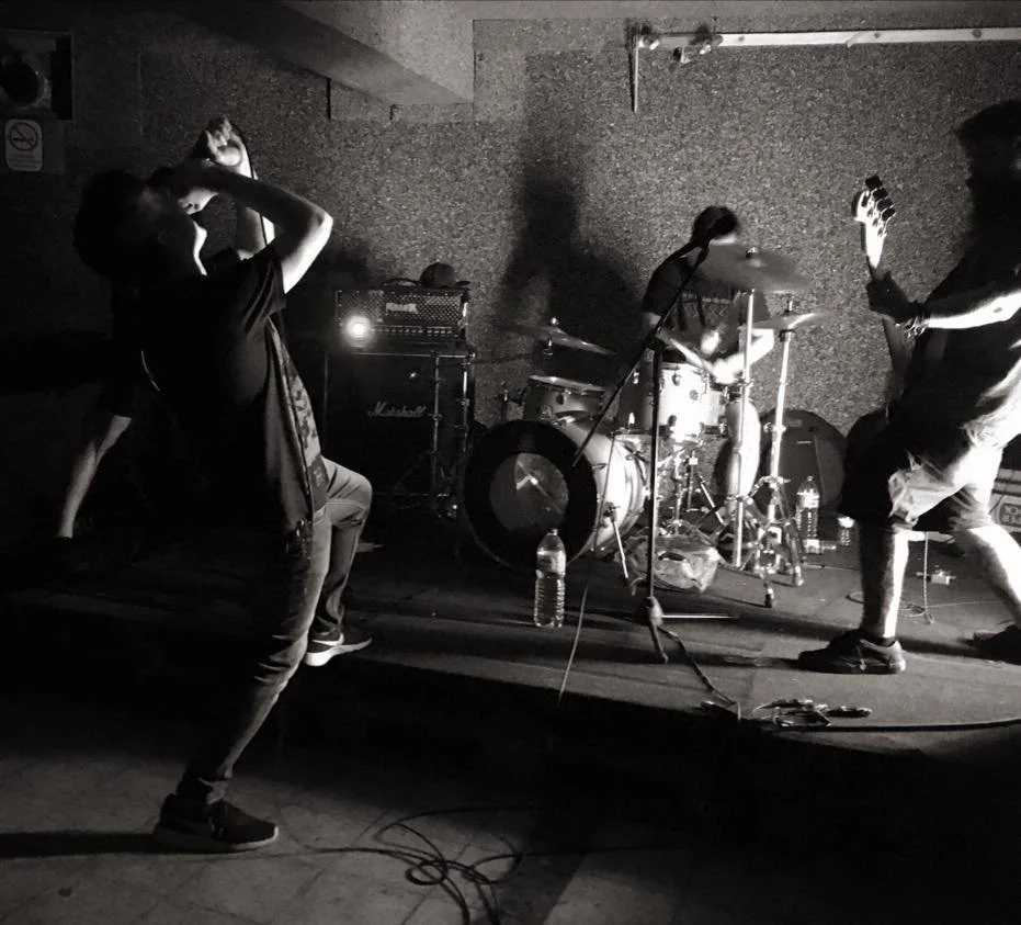Three musicians performing on stage: one holding a microphone and singing, another playing drums, and the third playing an electric guitar. The stage has amplifiers and water bottles.