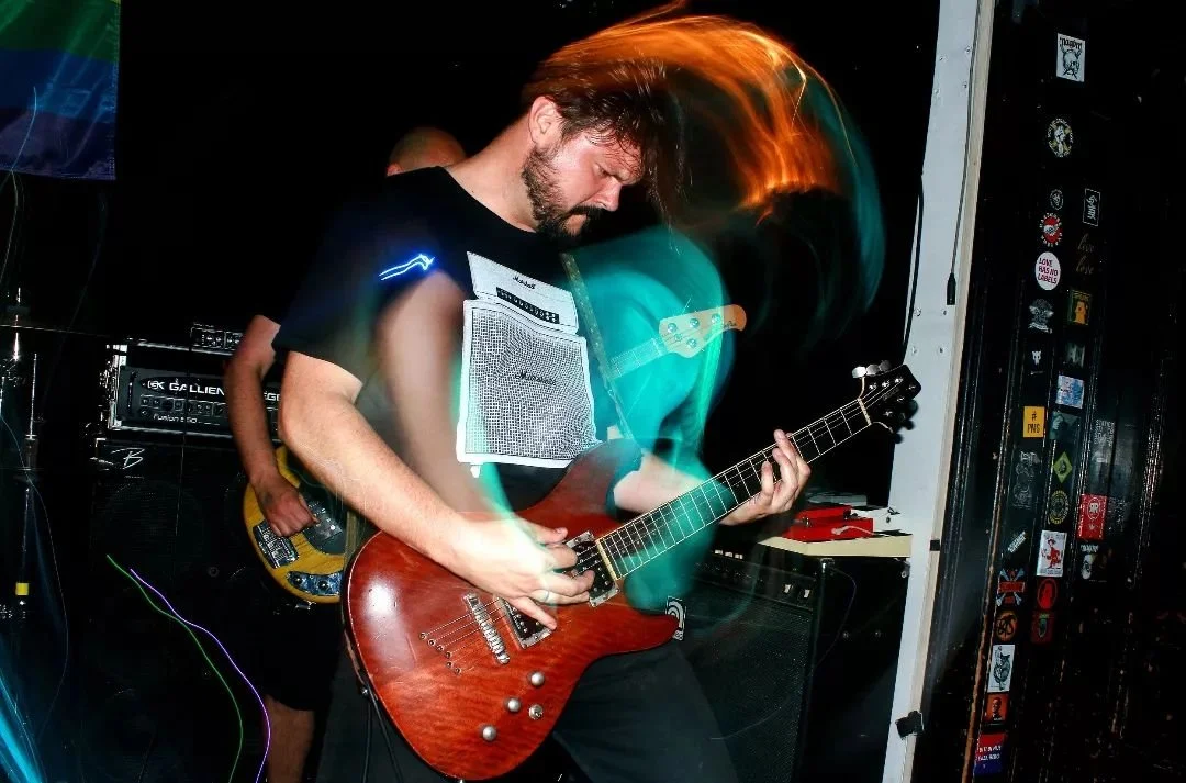 Musician playing an electric guitar with colorful light streaks, in a dimly lit venue with musical equipment and stickers on the wall.