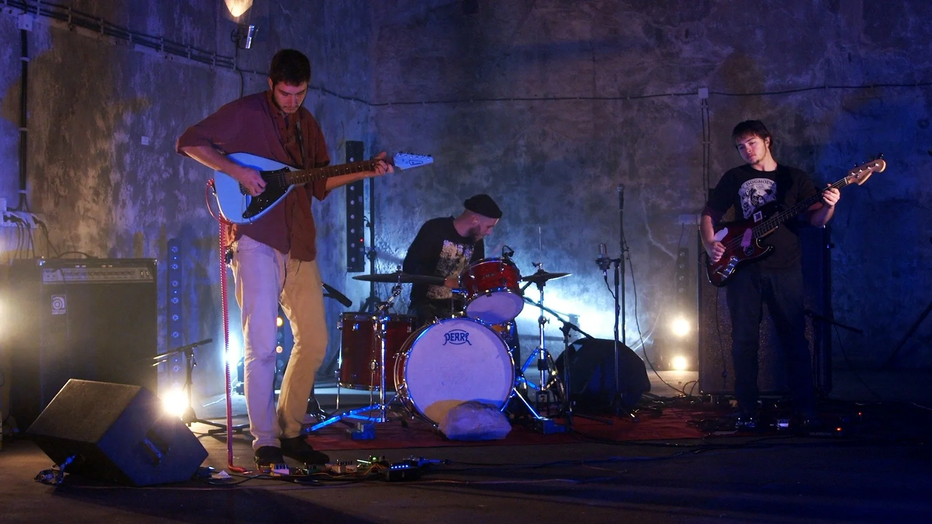 A band performing live on stage with three male musicians: one playing an electric guitar, one on drums, and one playing a bass guitar. The stage has low lighting with blue and purple hues.