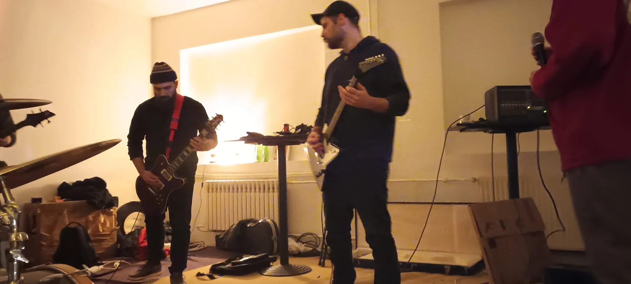 Musicians practicing in a dimly lit room, with guitars and audio equipment around, including a person in a red hoodie holding a microphone.