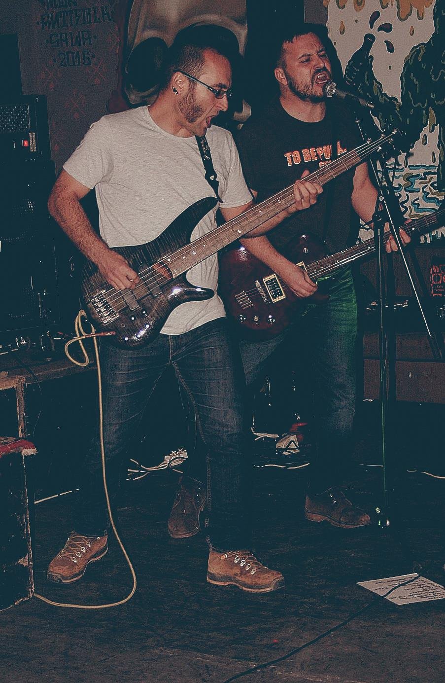 Two male musicians performing with electric guitars and singing into a microphone in a dimly lit venue.
