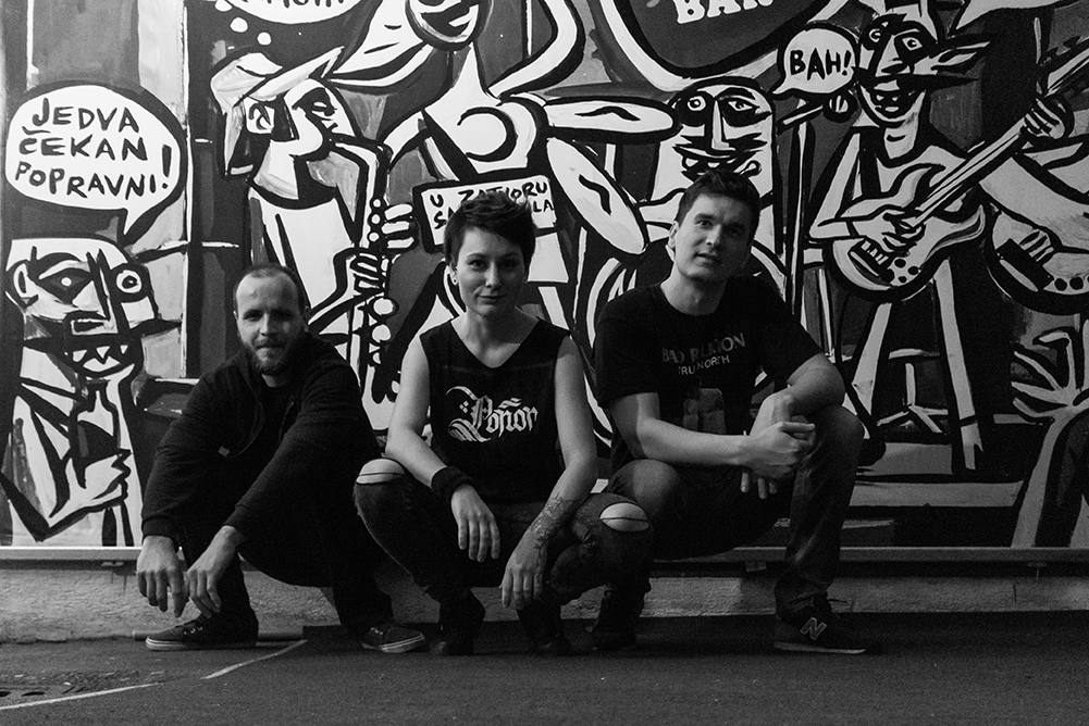 Three people crouching in front of a black and white graffiti mural, featuring stylized cartoon-like characters and speech bubbles.