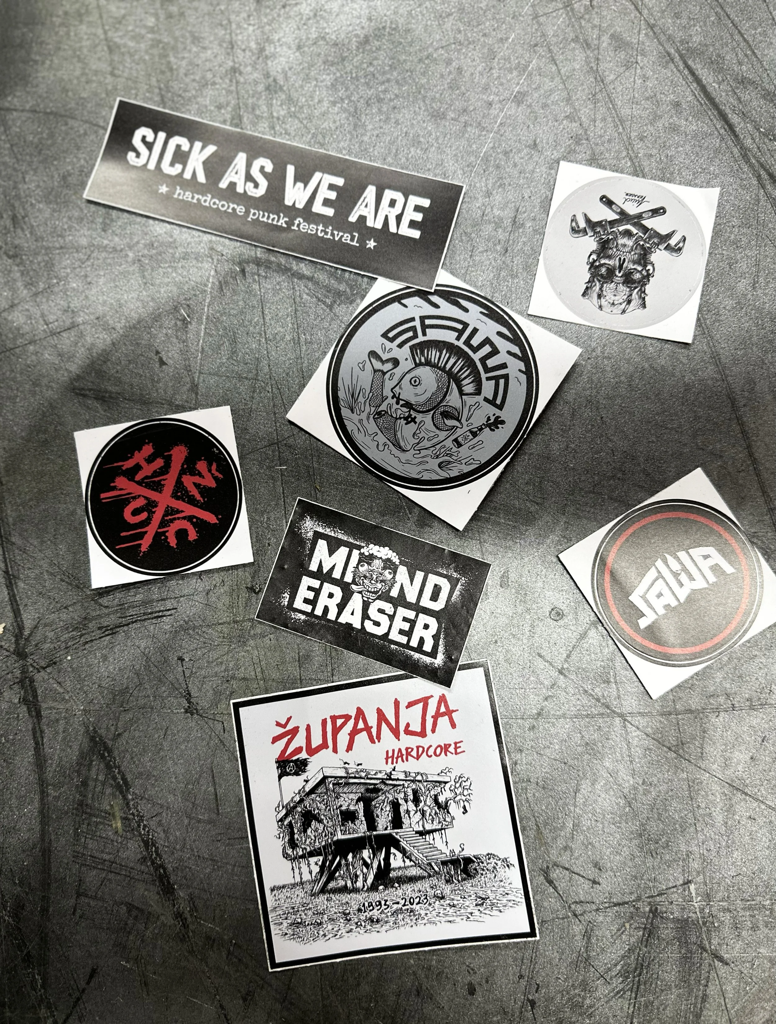 A collection of black and white stickers on a scratched metal surface. The stickers include phrases such as 'SICK AS WE ARE' with 'hardcore punk festival,' and images like a fish surrounded by various objects, and a building with the years 1993-2023.