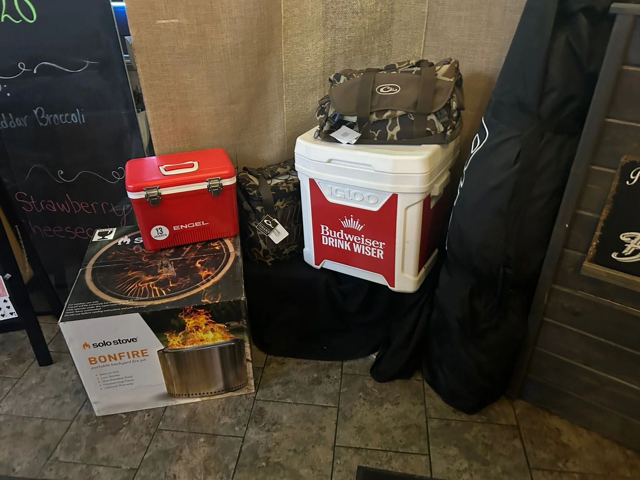 Indoor scene showing a blackboard, a red portable fire pit, a white cooler with a Budweiser sign, and various bags and clothing items stacked in a corner.
