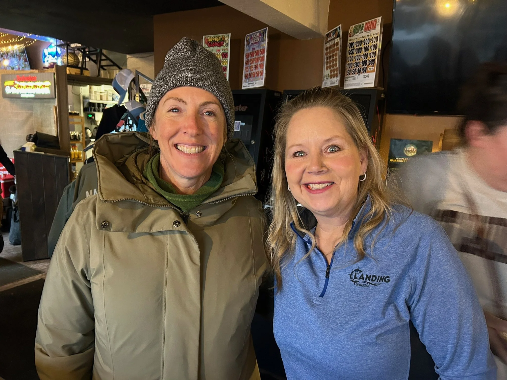 Two women smiling and posing for a photo inside a bar or restaurant. One woman is wearing a beanie and a beige jacket, and the other woman is wearing a blue hoodie with a logo. The background shows menu boards, a television screen, and some blurred f