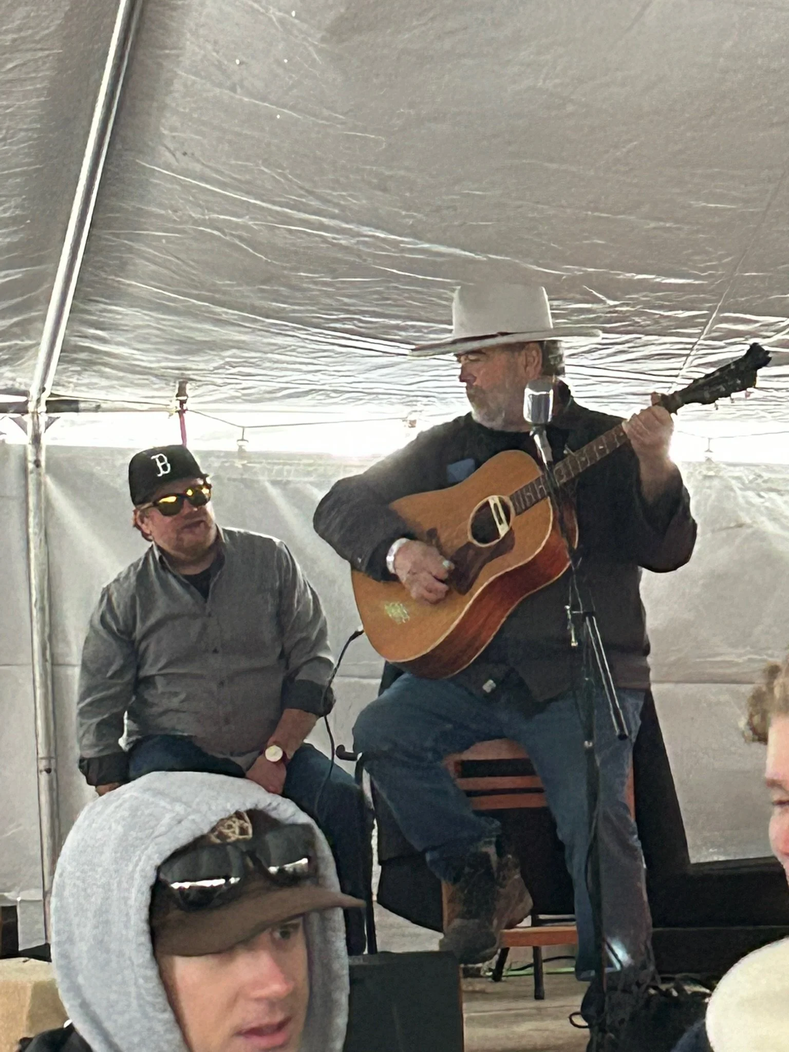 A man wearing a white cowboy hat, black jacket, and glasses playing an acoustic guitar under a large tent. Another man in a black baseball cap with a letter 'B' and sunglasses is seated nearby, listening.