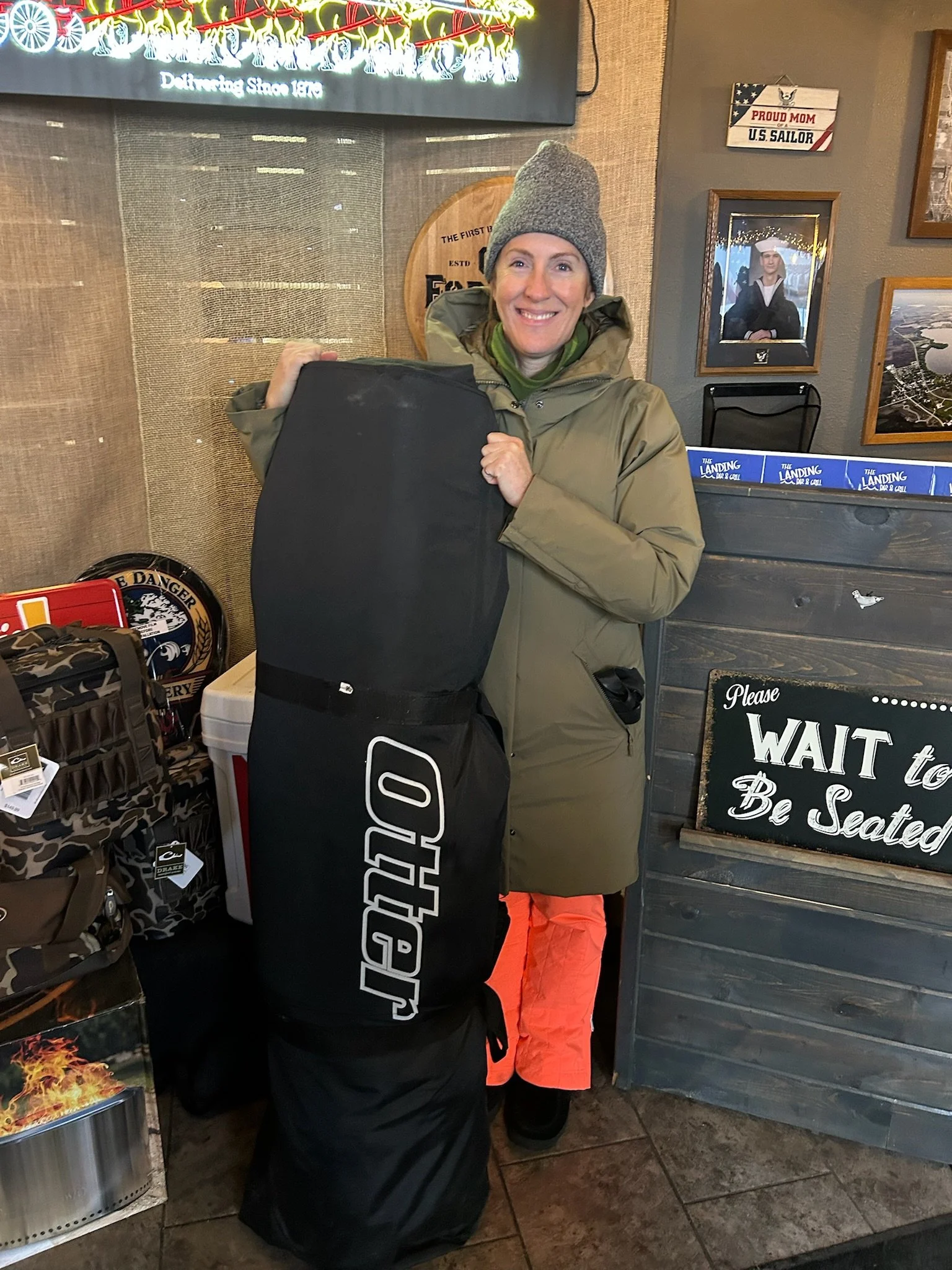 A woman dressed in outdoor clothing, including a gray knit hat and a tan jacket, smiling and standing inside a store. She is holding a large black item, possibly a bag or case. The background features wooden wall decor, framed photos, and a sign that