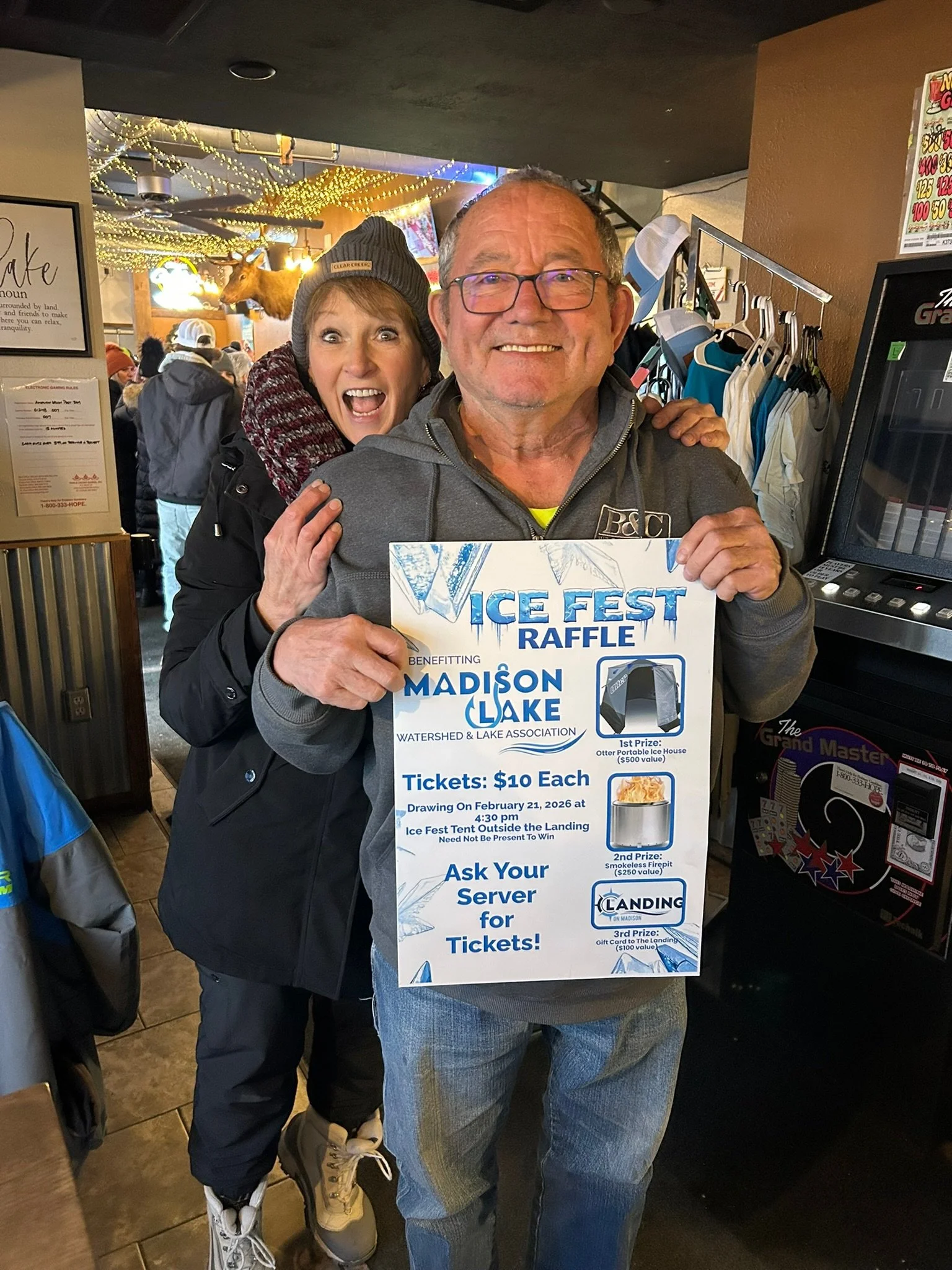 Two smiling people, a woman and a man, holding a sign for an ice fest raffle. The woman is behind the man, wearing a beanie and smiling widely. The man is in front, wearing glasses and a gray jacket, holding the raffle sign which mentions benefits fo
