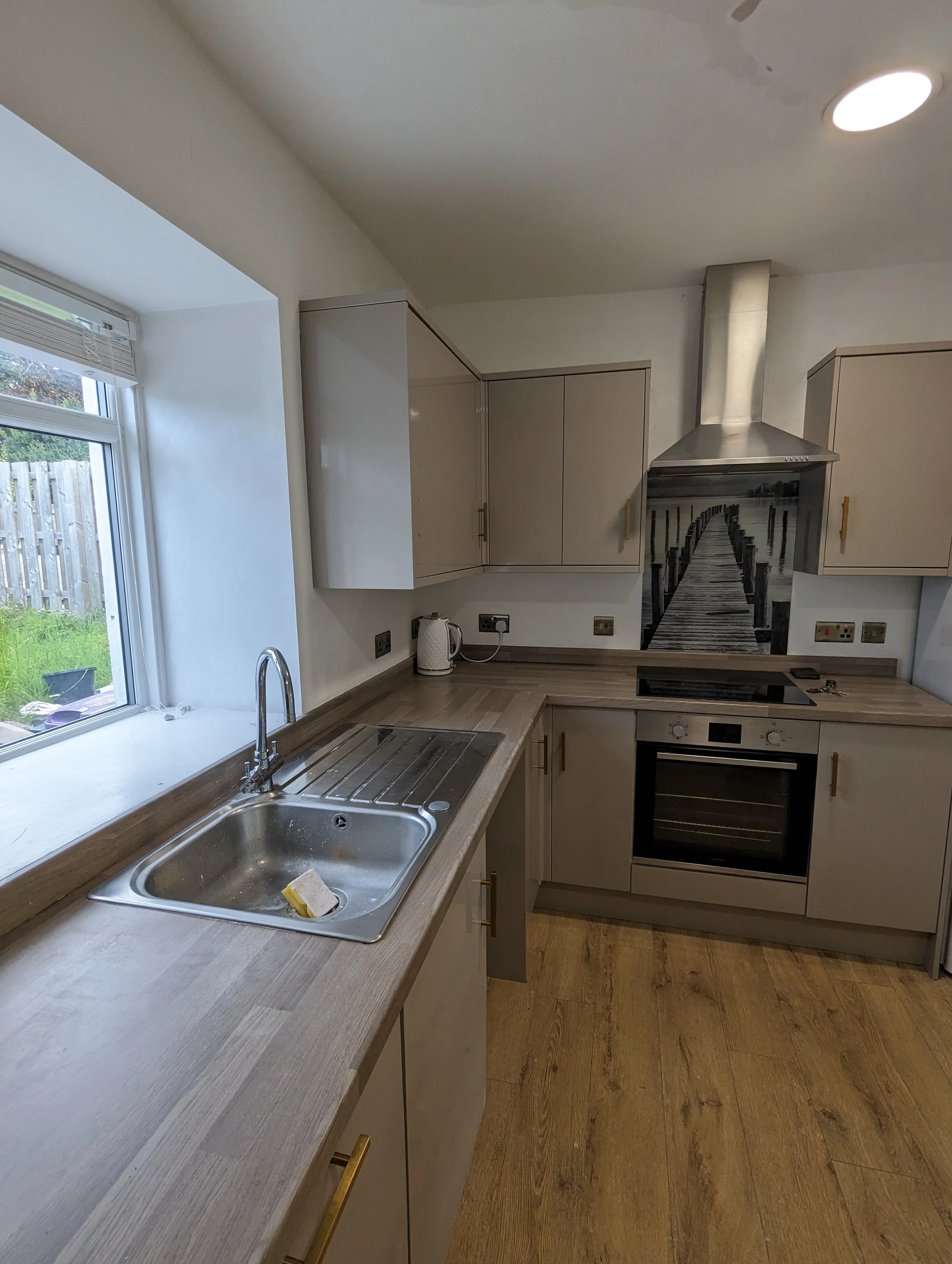 A modern kitchen with beige cabinets, a stainless steel oven, a stove, and a range hood featuring a painted pier scene. There is a window over the sink showing a backyard with grass and a wooden fence.