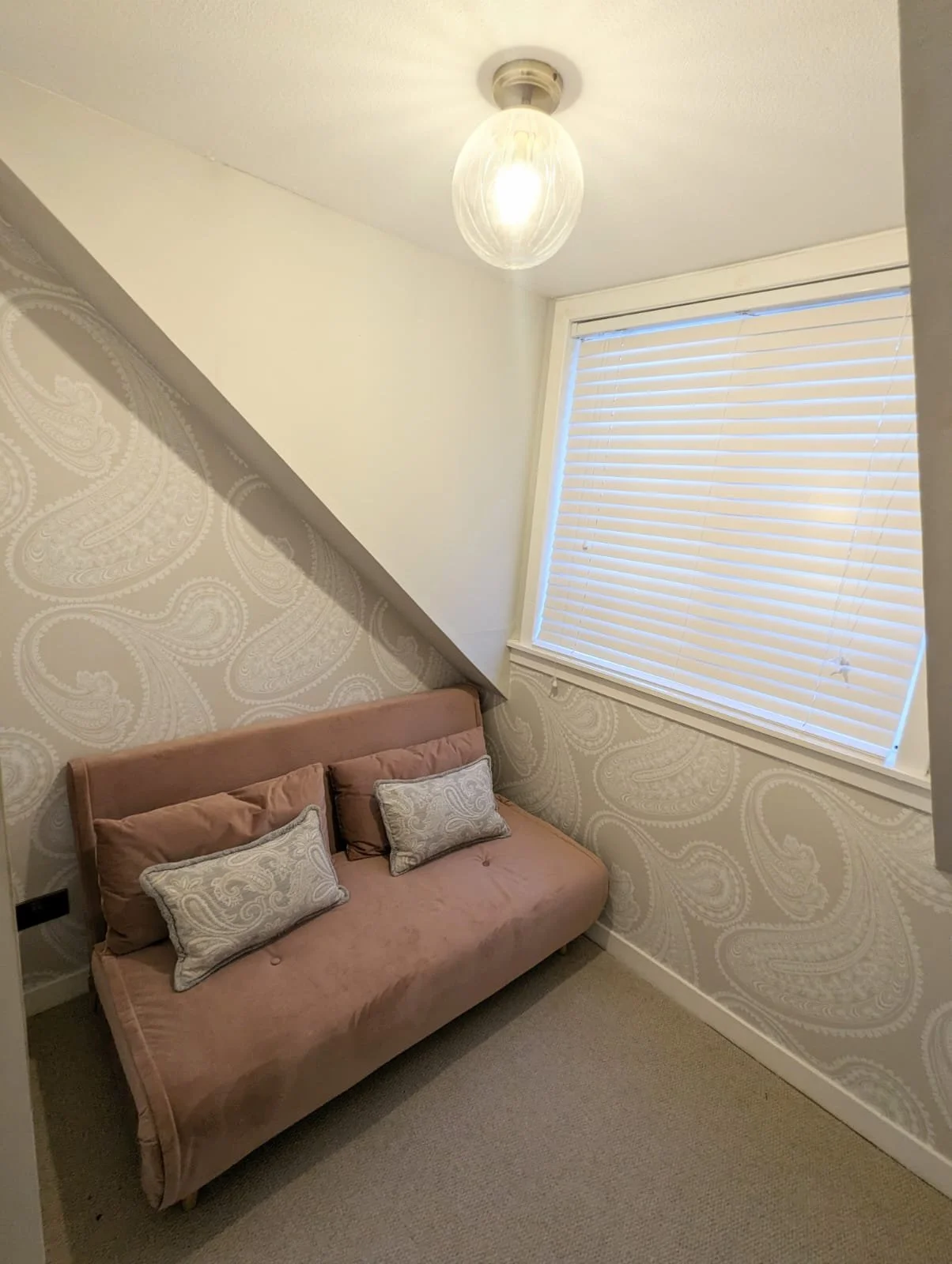 Pink velvet sofa with two pillows and one bolster pillow, against a patterned wallpapered wall, beneath a sloped ceiling, near a window with closed blinds, in a small room with beige carpet and a ceiling light fixture.