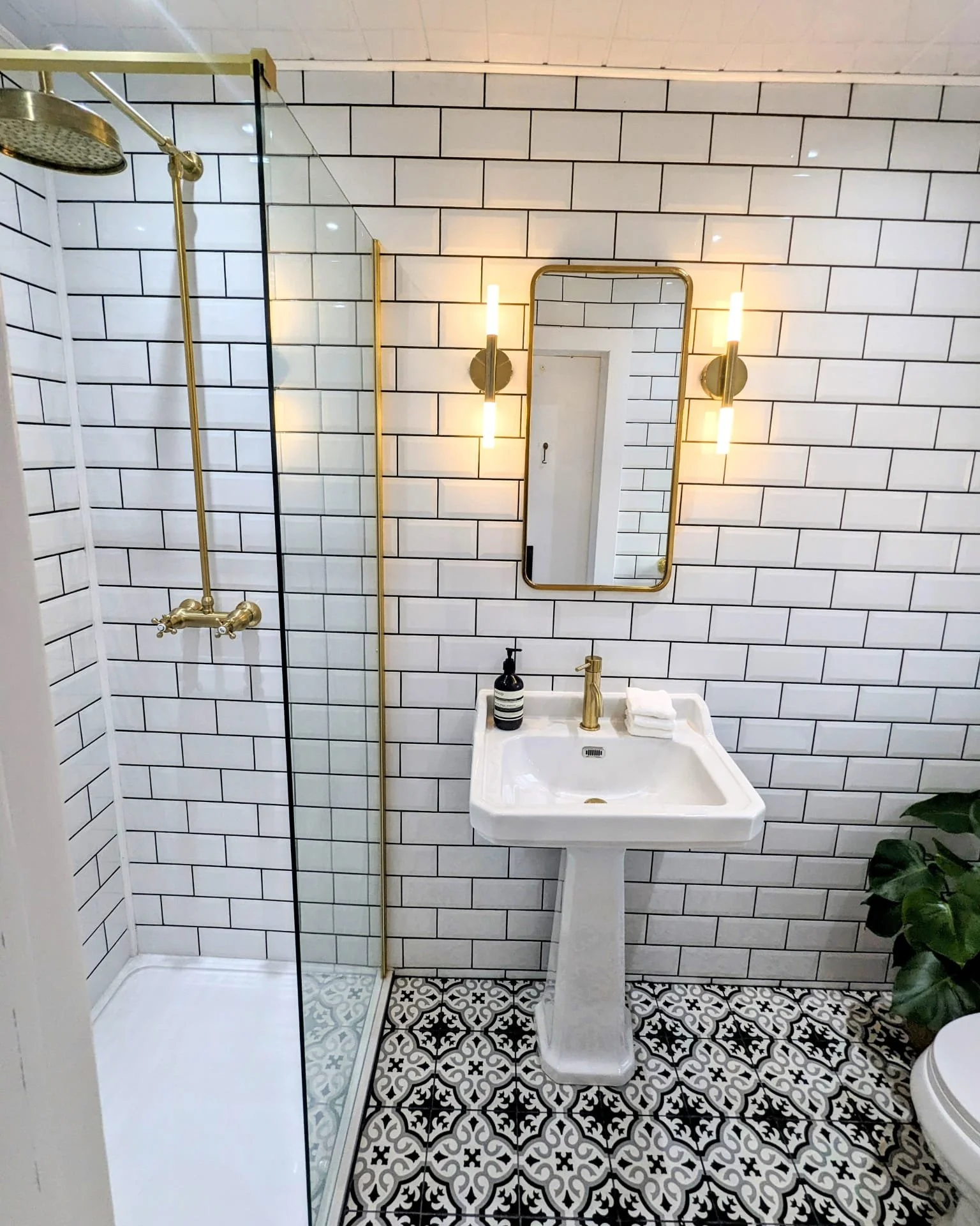 Modern bathroom with white subway tiles, black grout, black and white patterned floor, gold fixtures, rectangular mirror, and a small white pedestal sink with soap and towels. A rain shower head is in the corner.