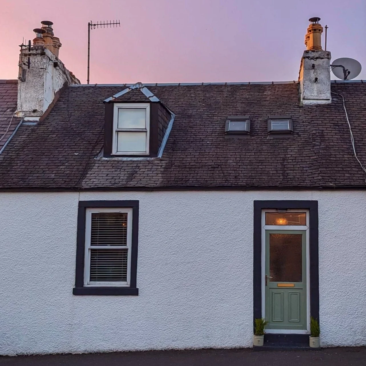 Front of a white house with black trim, two windows with blinds, a green front door with a mail slot, and potted plants on either side. The upper roof has one dormer window and two skylights, with chimneys and antennas visible against a pink sky.