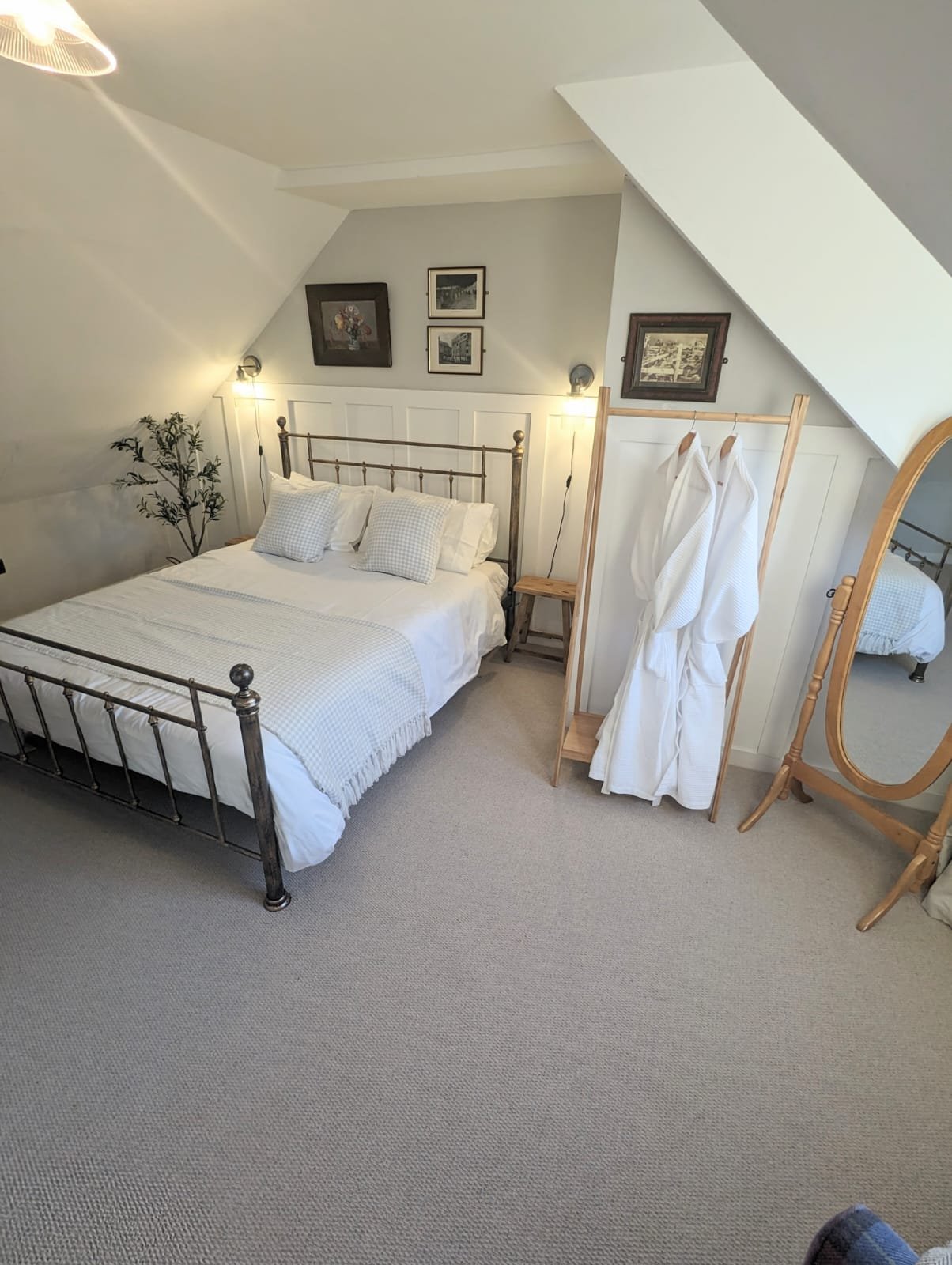 A cozy bedroom with a white bed, pillows, and a metal bed frame. There is a wooden bench, a standing mirror, and a robe on a wooden rack. Walls feature framed pictures, and there are two wall-mounted lamps.