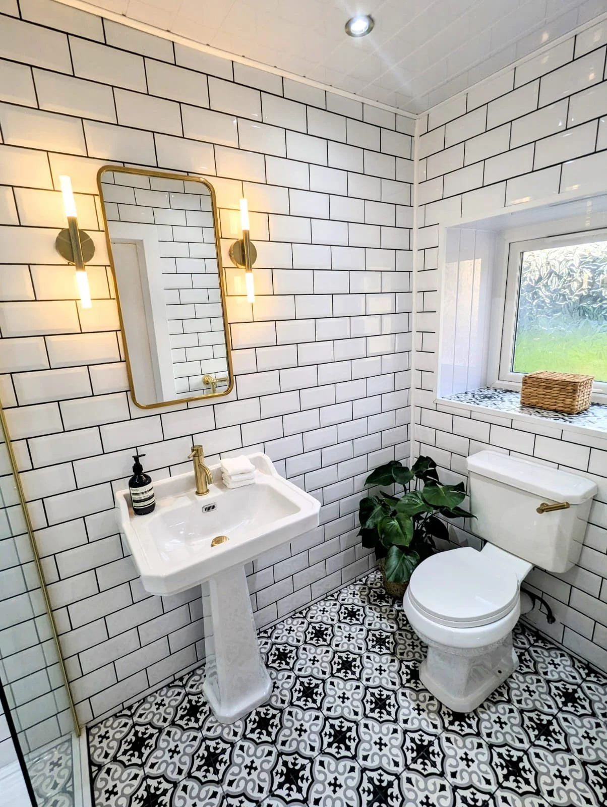 A bathroom with white subway tile walls, black grout lines, decorative black and white patterned floor tiles, a white pedestal sink with gold fixtures, a wall mirror with gold frame, wall-mounted gold lights, a toilet, a window with frosted glass, a wicker basket on the windowsill, and a potted green plant near the toilet.