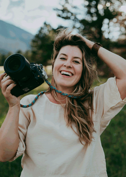 A woman smiling and holding a camera outdoors in a natural setting with trees and mountains in the background.