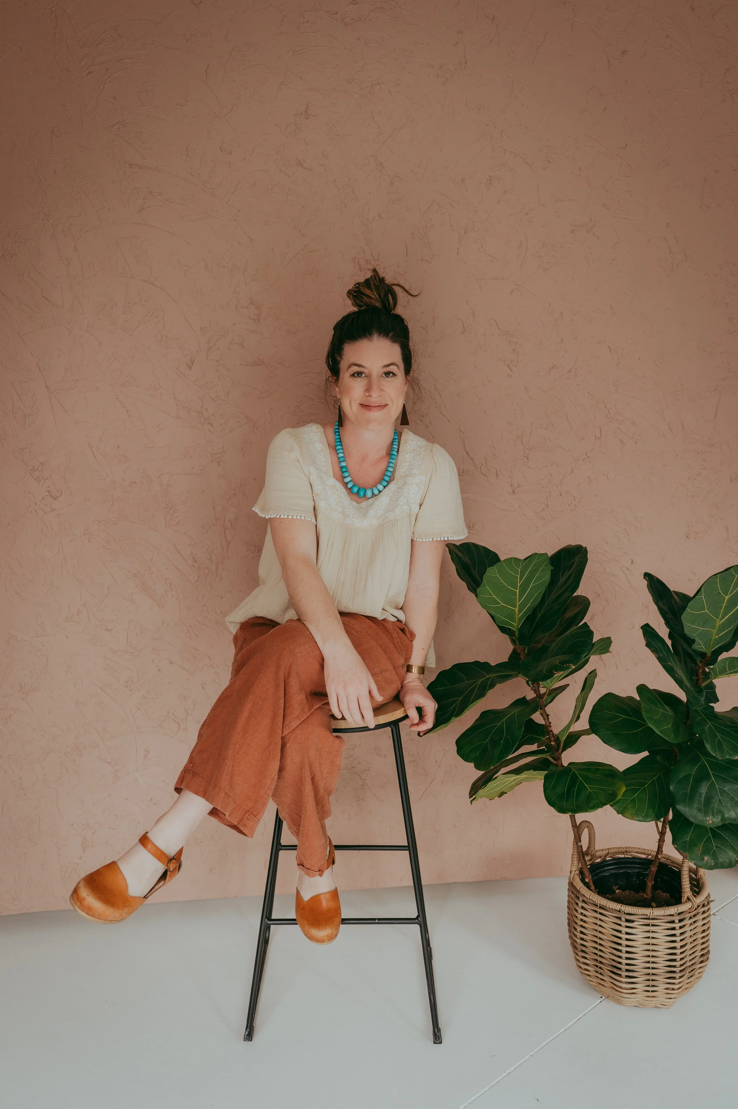 A woman with a top bun hairstyle, wearing a cream blouse, brown pants, and brown shoes, sitting on a stool beside a potted plant with large green leaves, against a pink wall background.