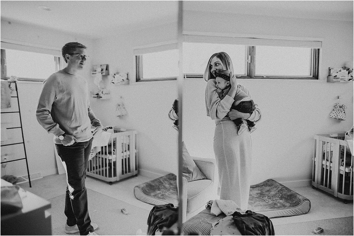 A woman holding a baby, standing in a bright room with windows, smiling at a man who is standing nearby. The room has cribs, toys, and a rug, appearing to be a nursery or playroom.