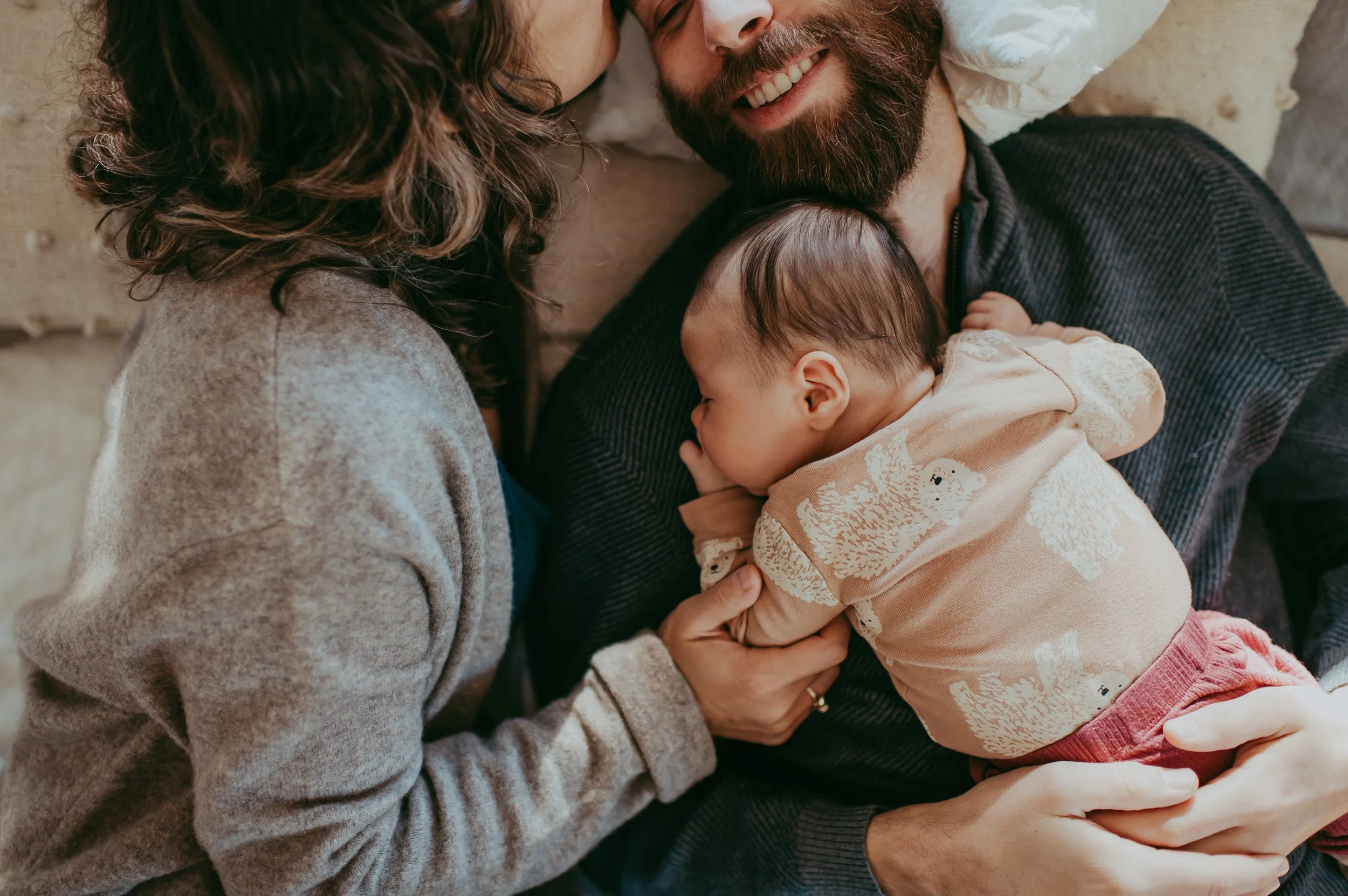 A family scene showing a man with a beard holding a young child, a woman with curly hair leaning in, all sharing a joyful moment together.