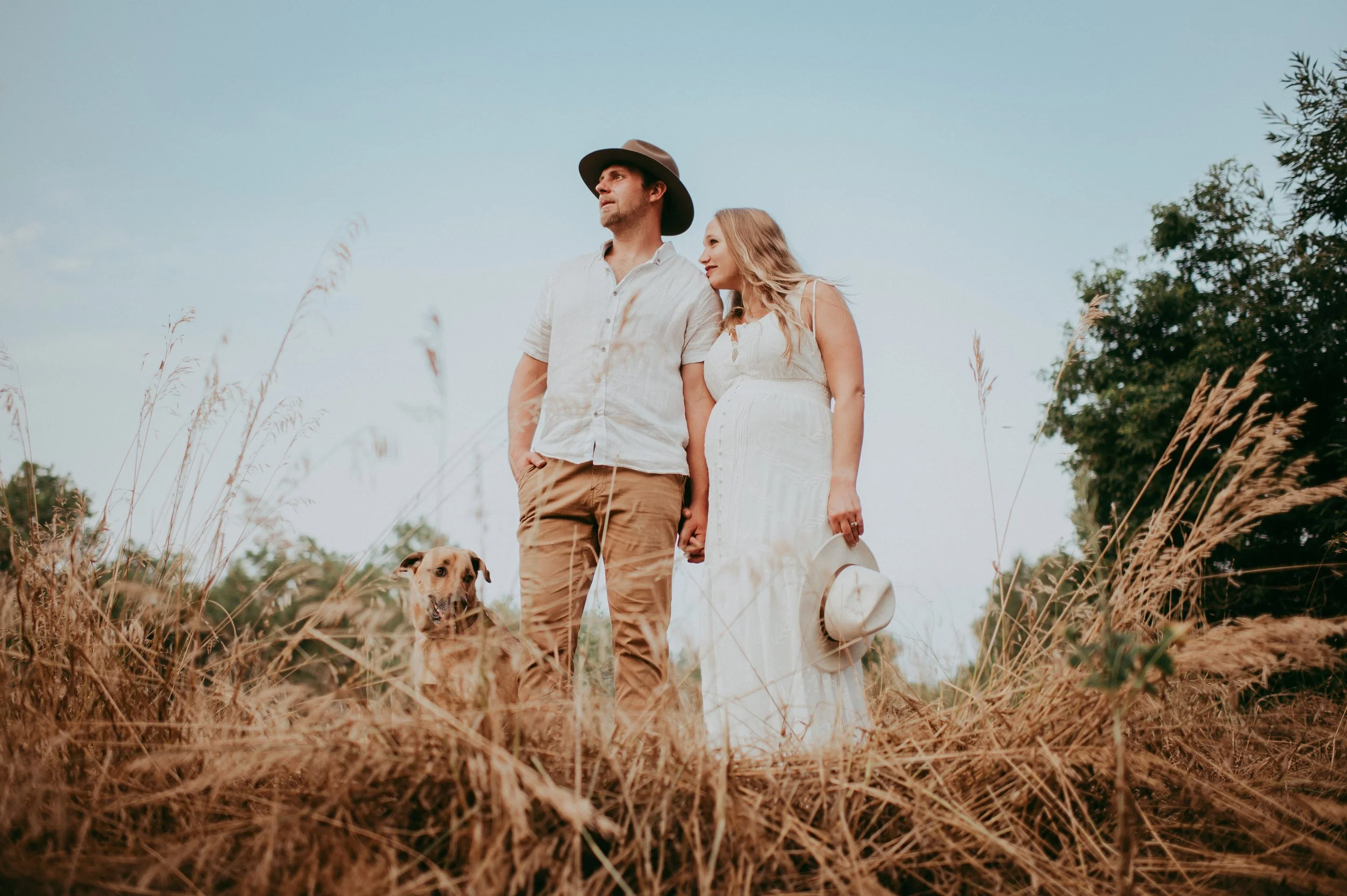 A couple holding hands with a dog in a field of tall dry grass during the daytime, with a clear sky and trees in the background.
