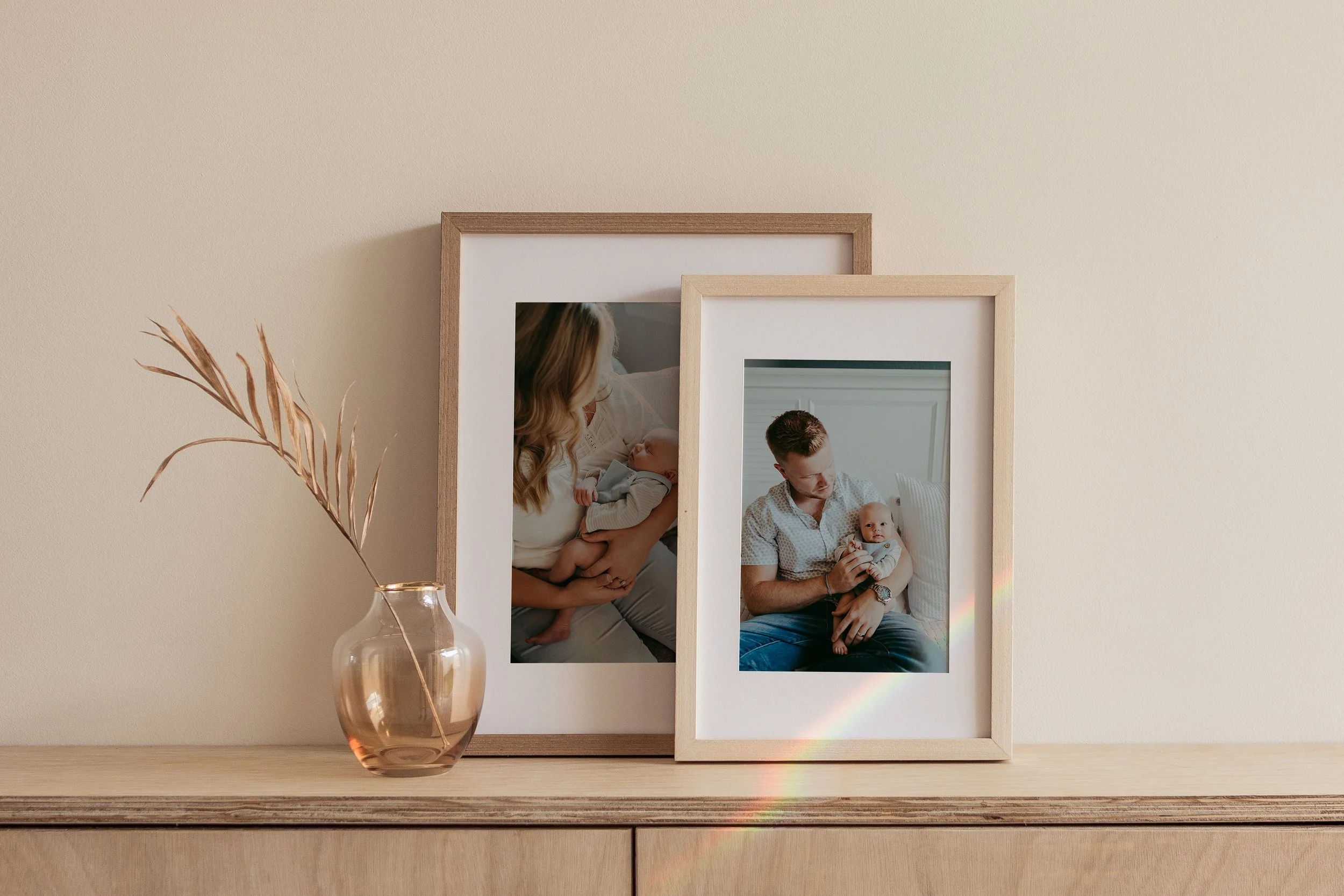A beige wall with two framed family photos, a small dried plant in a rose-colored glass vase, and sunlight creating a rainbow effect on the right side.