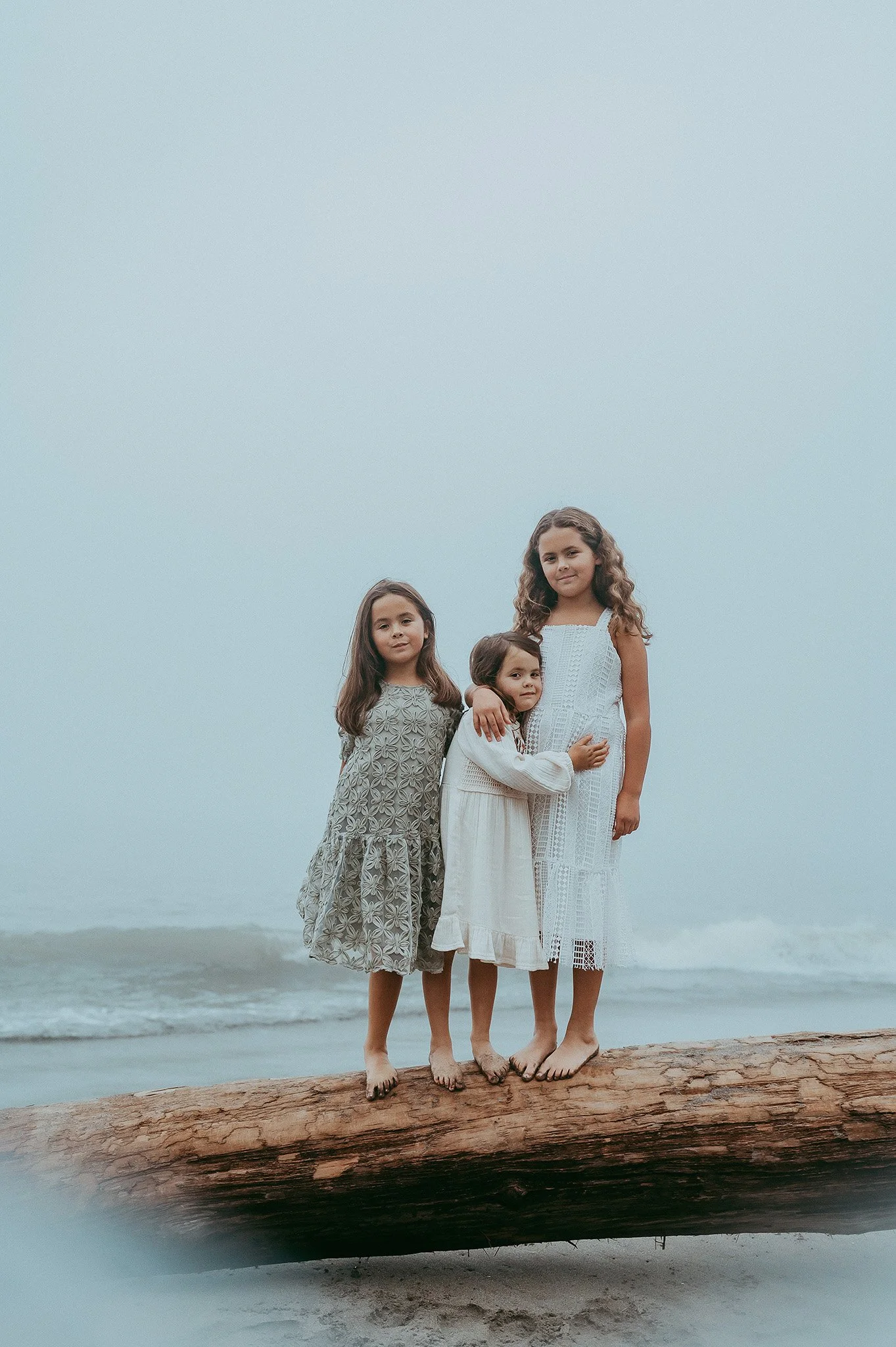 Four young girls in white and patterned dresses standing on a large fallen tree trunk on the beach by the water. Overcast sky in the background.