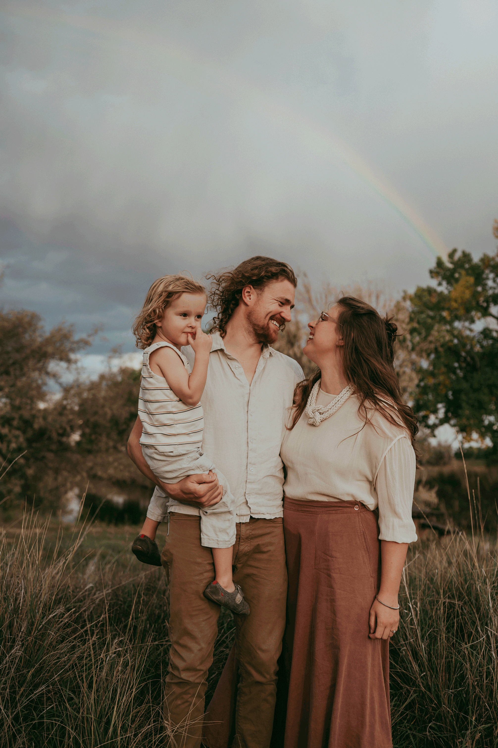 A family of three standing outdoors during daytime, with a rainbow in the cloudy sky behind them. The father is holding a young girl, while the mother looks at him and smiles.