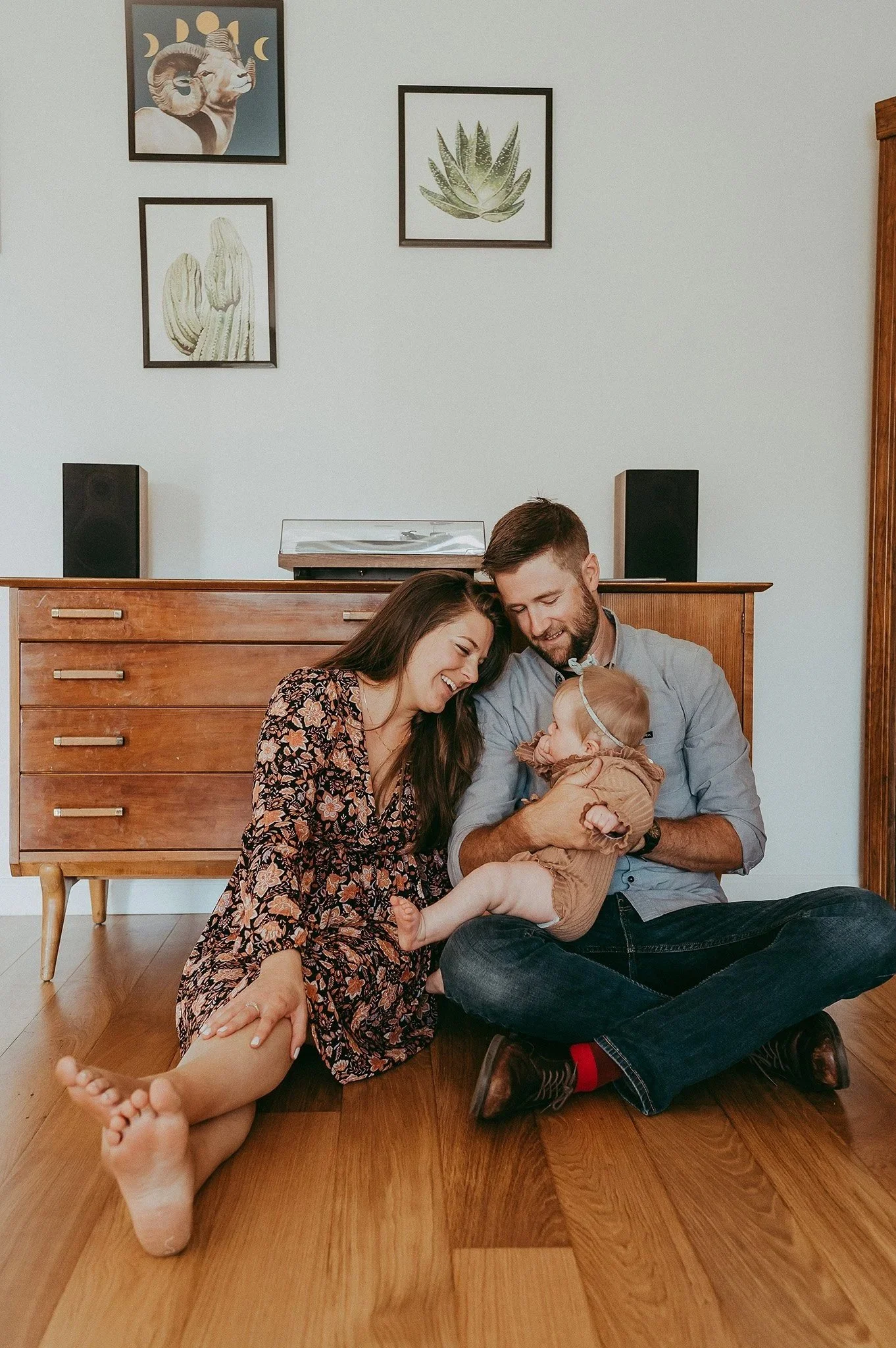 A family of three sitting on the wooden floor in a living room, smiling and playing with their baby girl. The mother wears a floral dress, the father is in a gray shirt and jeans, and the baby girl wears a brown outfit with a headband.