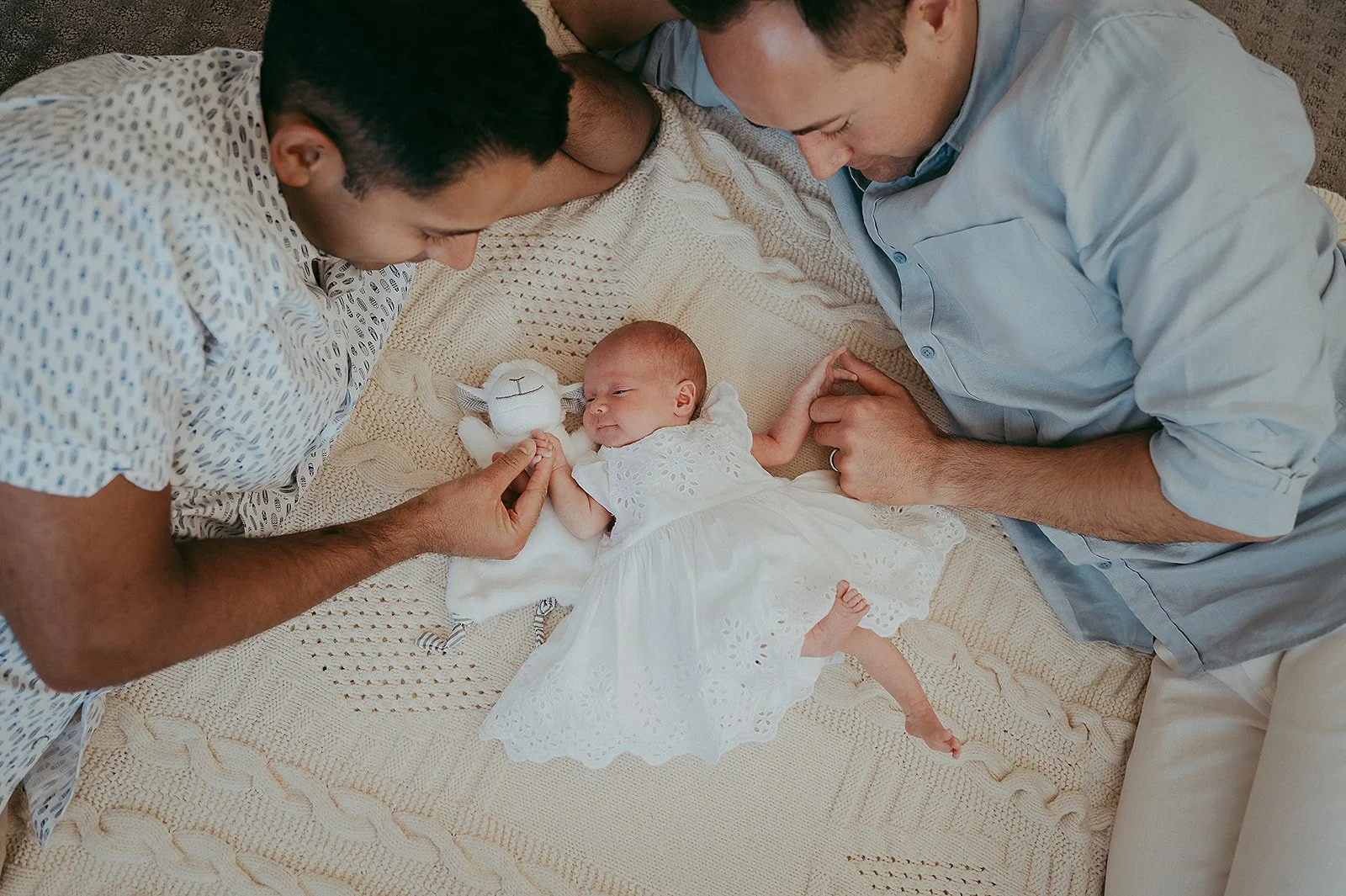 Two men lying on a blanket with a baby girl in a white dress between them, holding a small white stuffed animal.