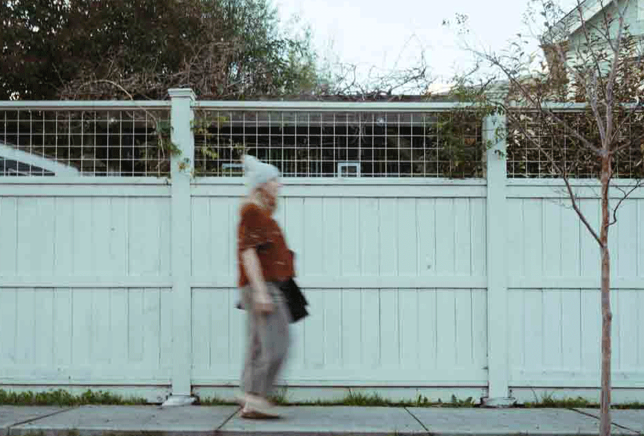 Older woman with white hair, wearing a brown top and gray pants, walking along a sidewalk next to a light-colored wooden fence with a small tree on the right and trees in the background.