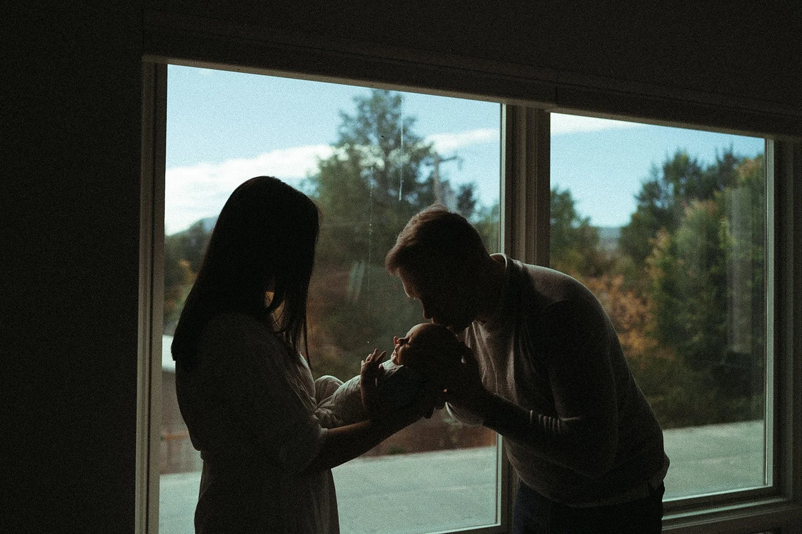 Silhouette of a family holding a newborn baby by a large window during daylight.
